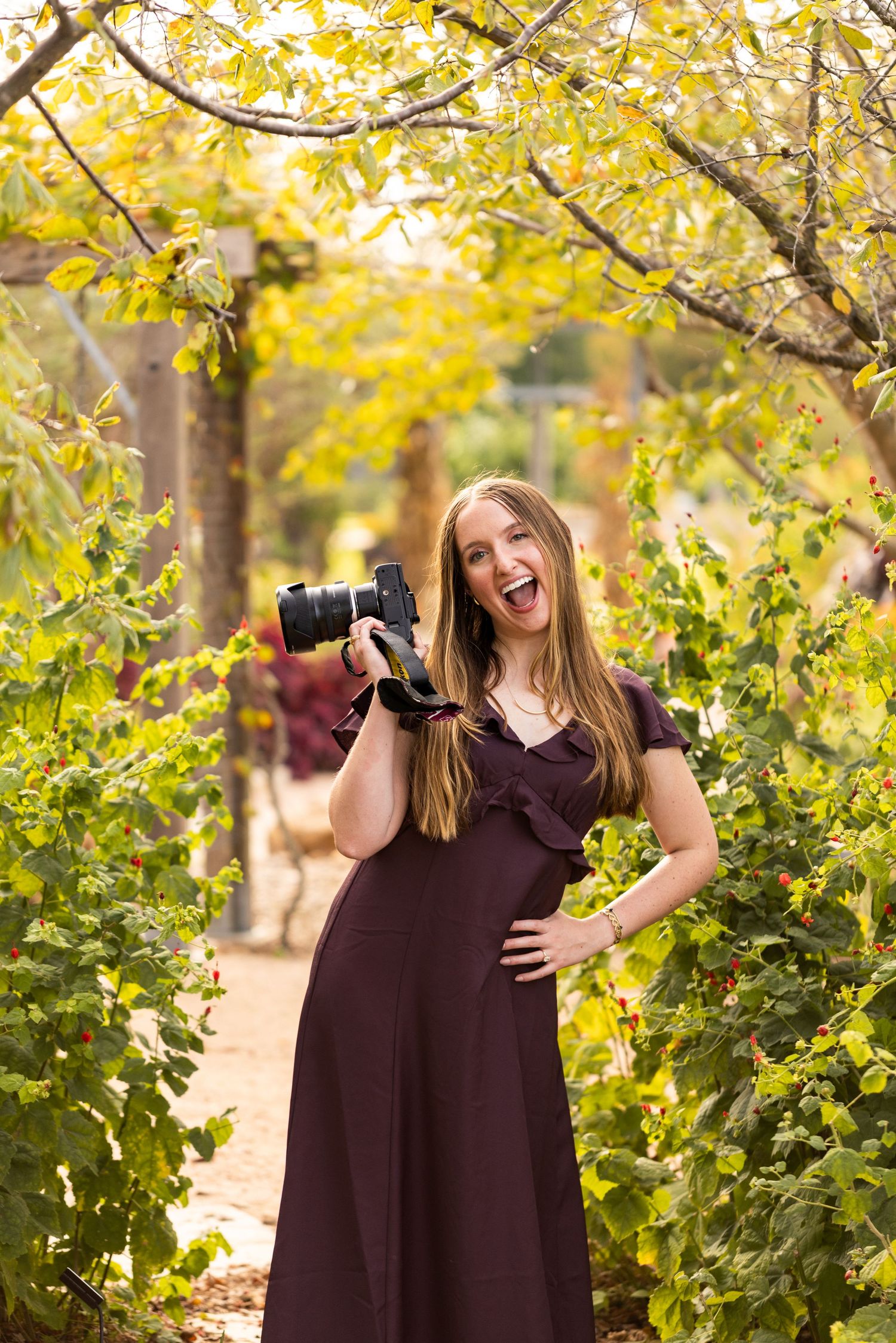 Smiling woman in a plum dress holds a DSLR camera on a garden path surrounded by lush greenery and golden autumn foliage.