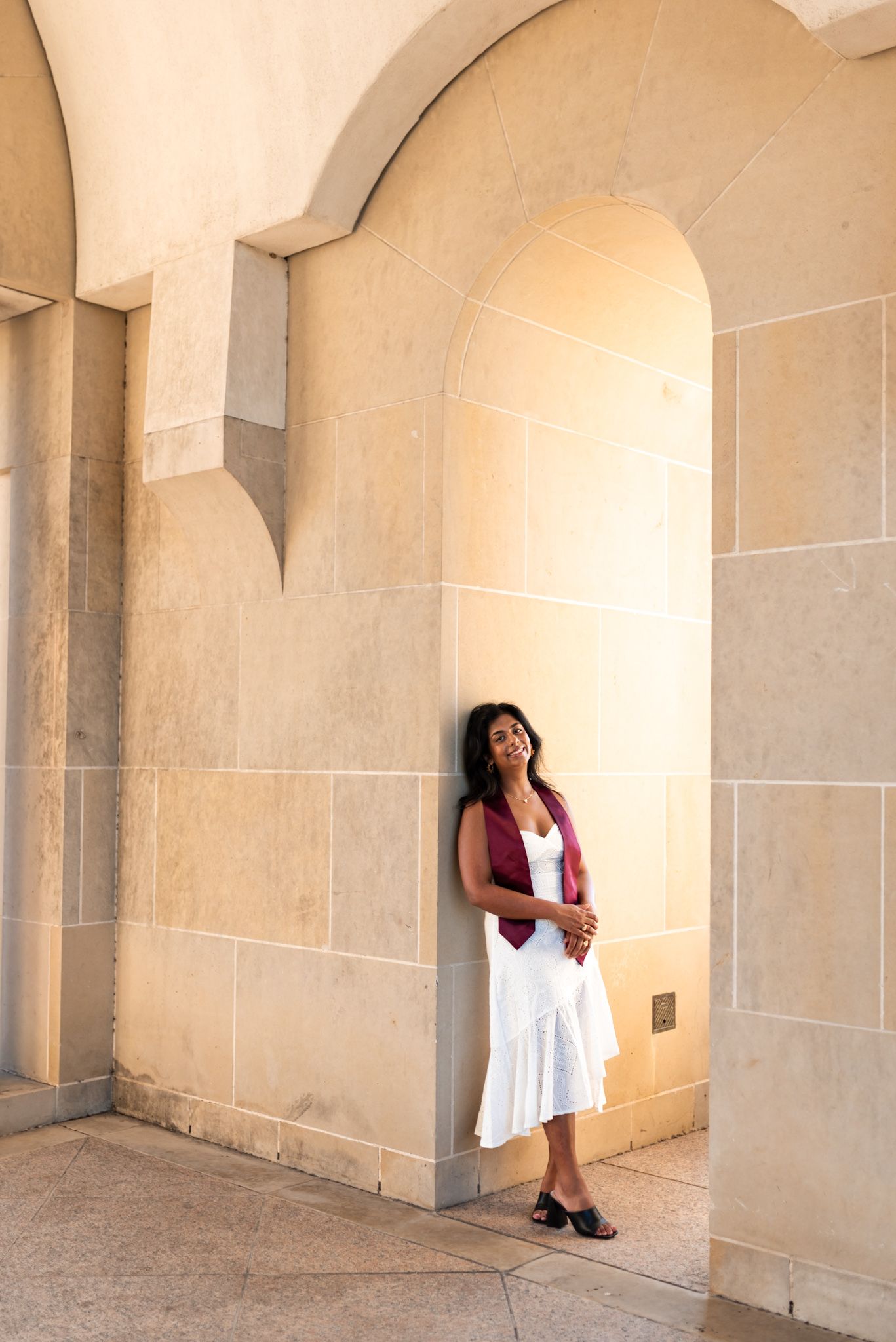 Graduate in white dress and maroon stole leaning against stone arch wall smiling.
