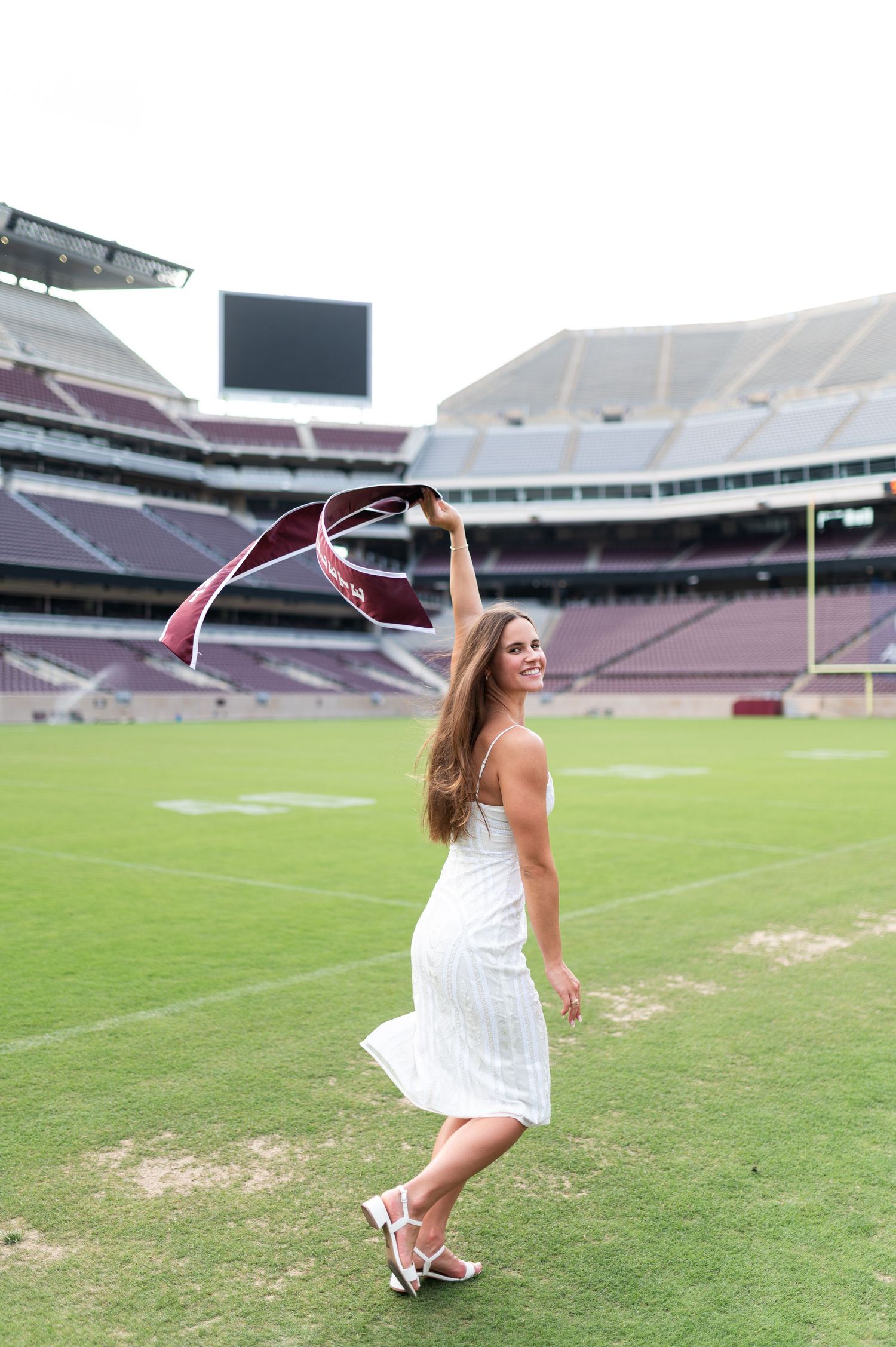 Female Texas A&M graduate in white dress waves maroon sash while walking on Kyle Field football turf.