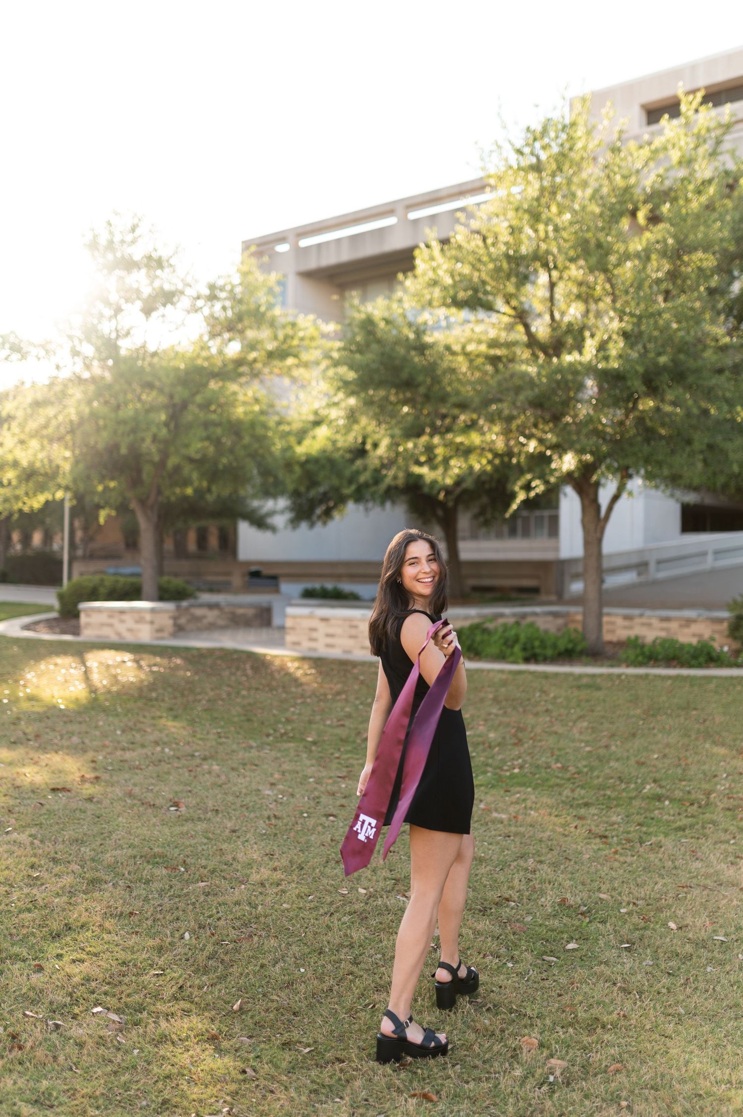 Texas A&M female graduate twirls maroon stole on sunny campus lawn at golden hour.