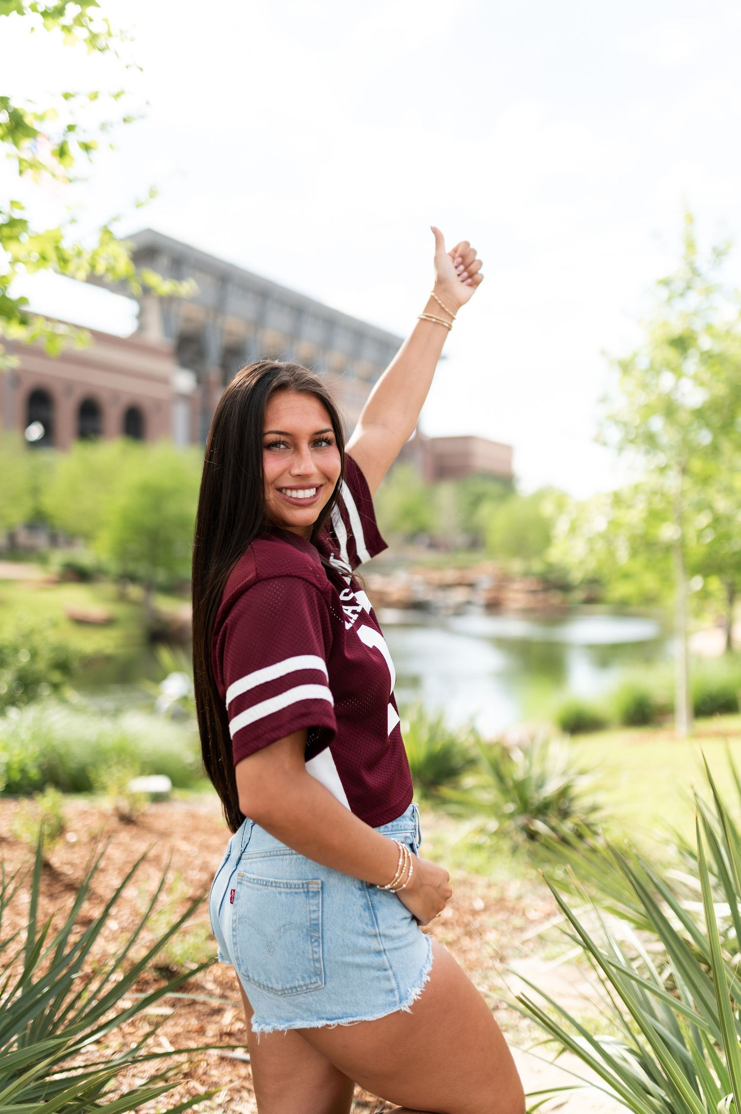 A smiling young woman in a Texas A&M jersey giving a thumbs up in a campus garden.