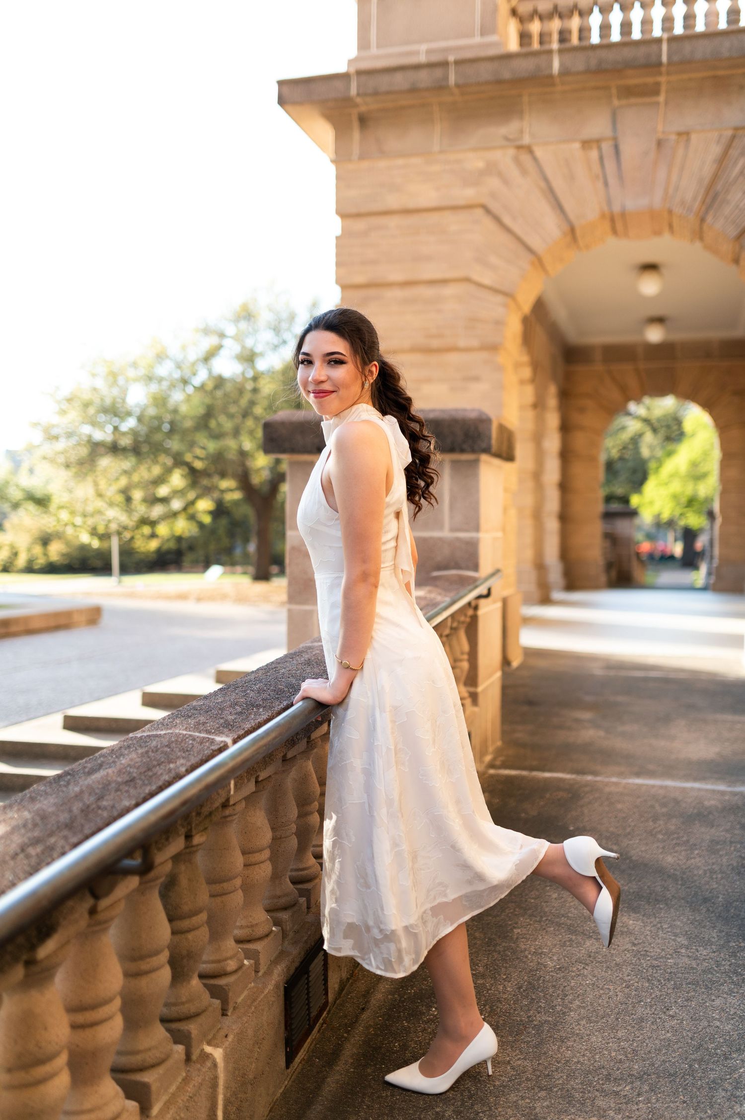 Young woman in white midi dress and heels poses gracefully on stone balcony of historic building.