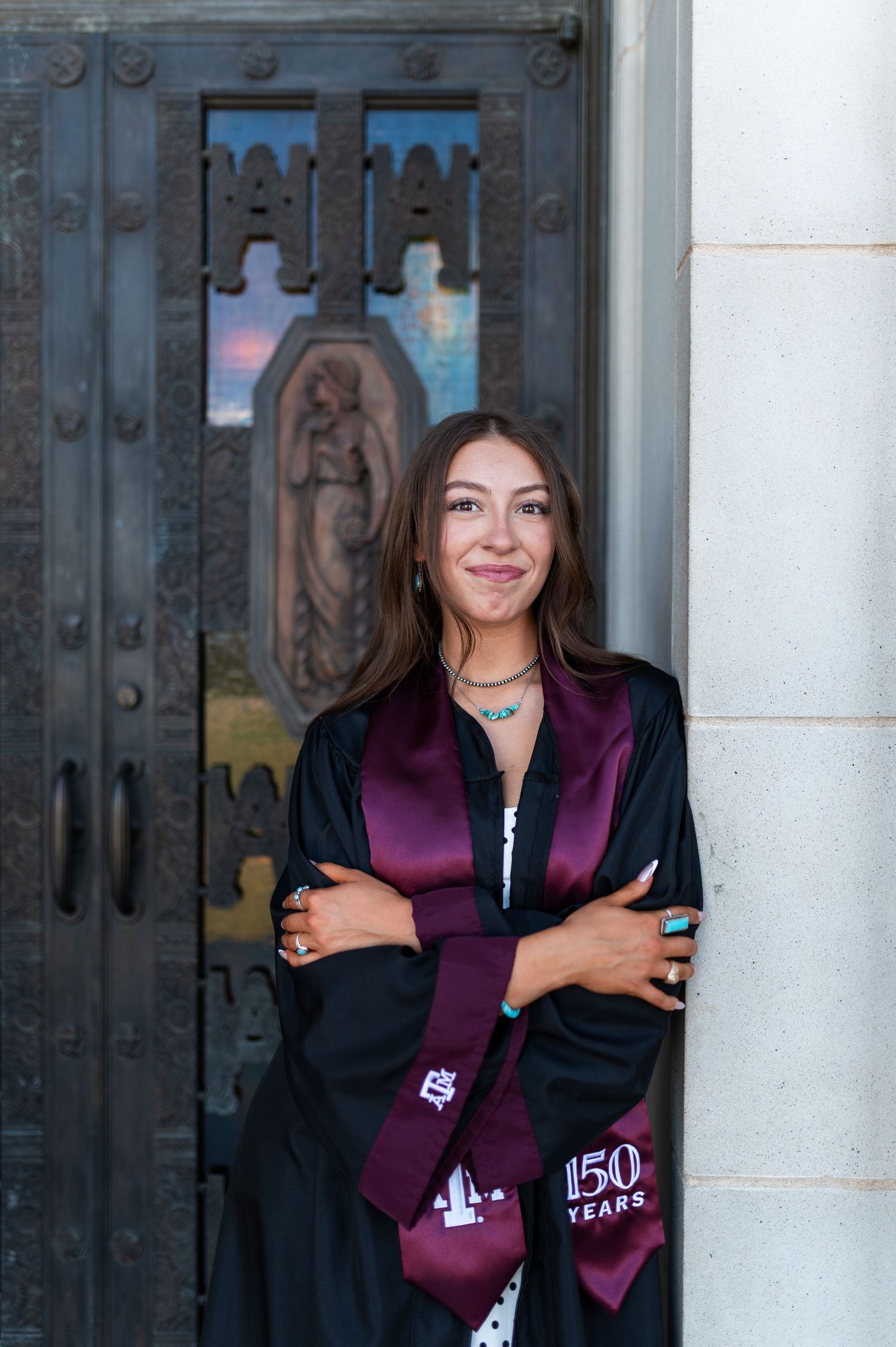 Smiling Texas A&M graduate poses with crossed arms in black graduation robe and maroon stole by ornate door. Grad Photos at the Admin
