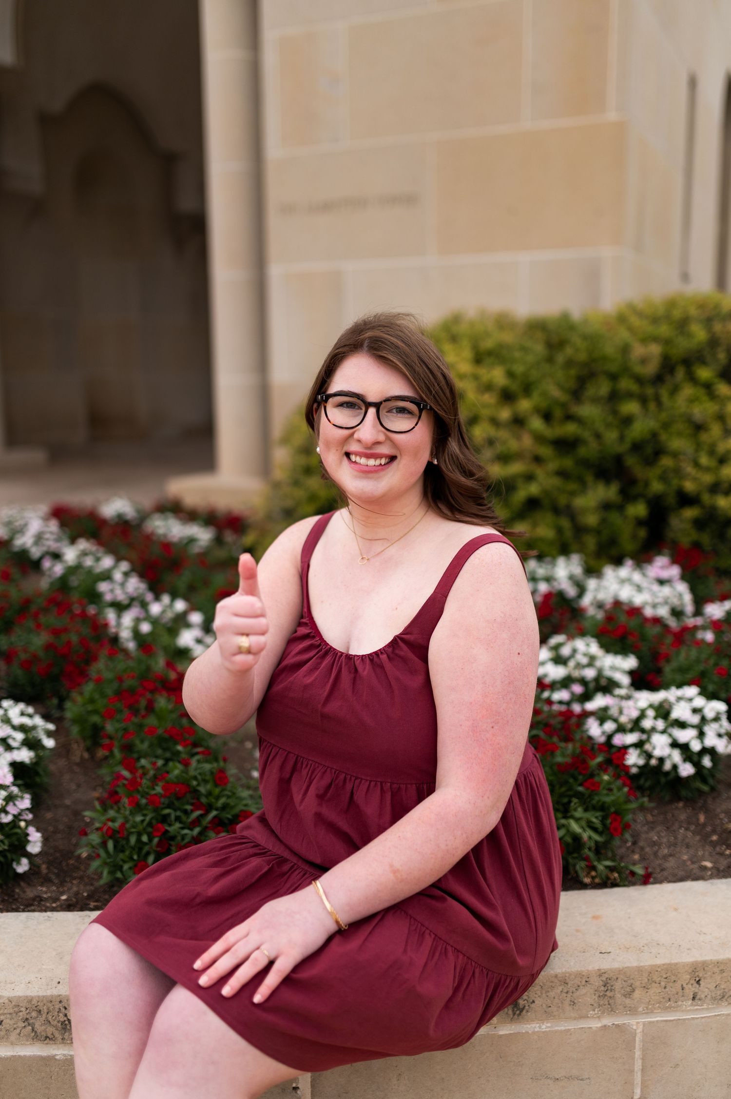 Texas A&M graduate in maroon dress giving thumbs up while seated near red and white flowers.