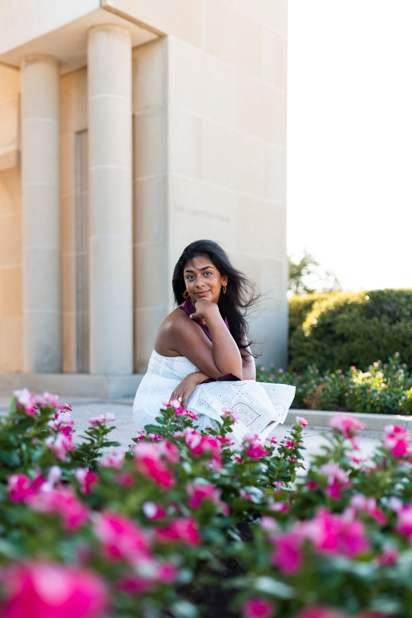 Young woman in white dress sitting near pink flowers by stone building columns.