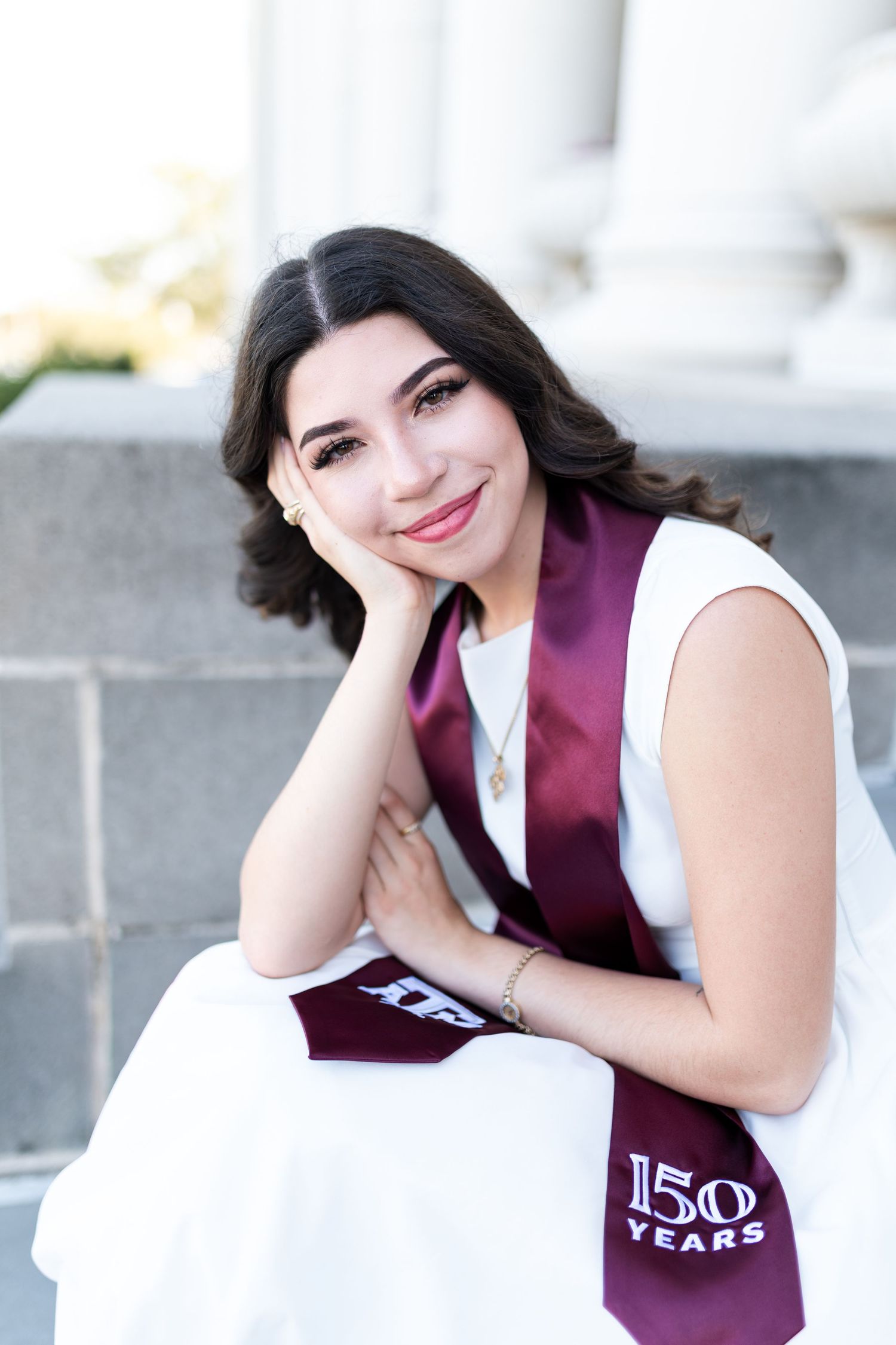 Smiling graduate in white dress with maroon Texas A&M 150 Years stole sitting on steps. Grad Photos at the Admin building