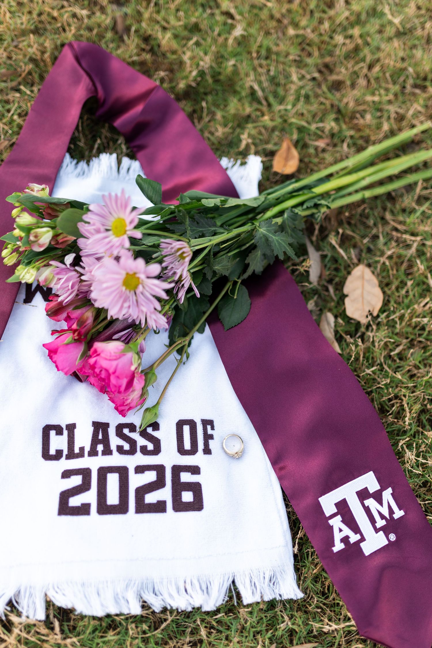 Texas A&M Class of 2026 white stole and maroon ATM sash with pink flower bouquet laying on grass.