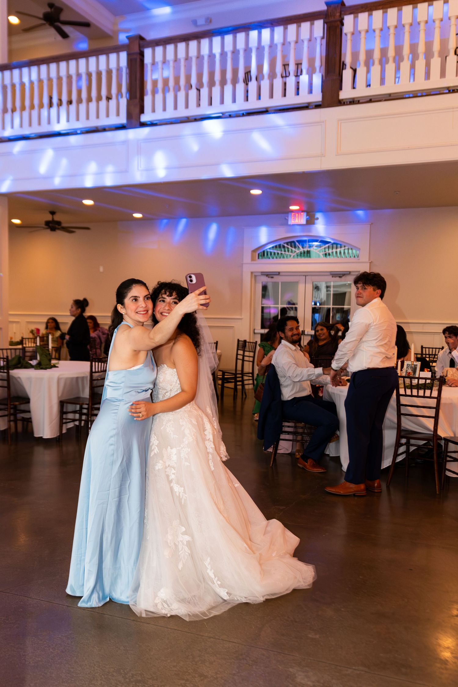Bride in lace gown taking selfie with bridesmaid in blue dress on dance floor at wedding reception.