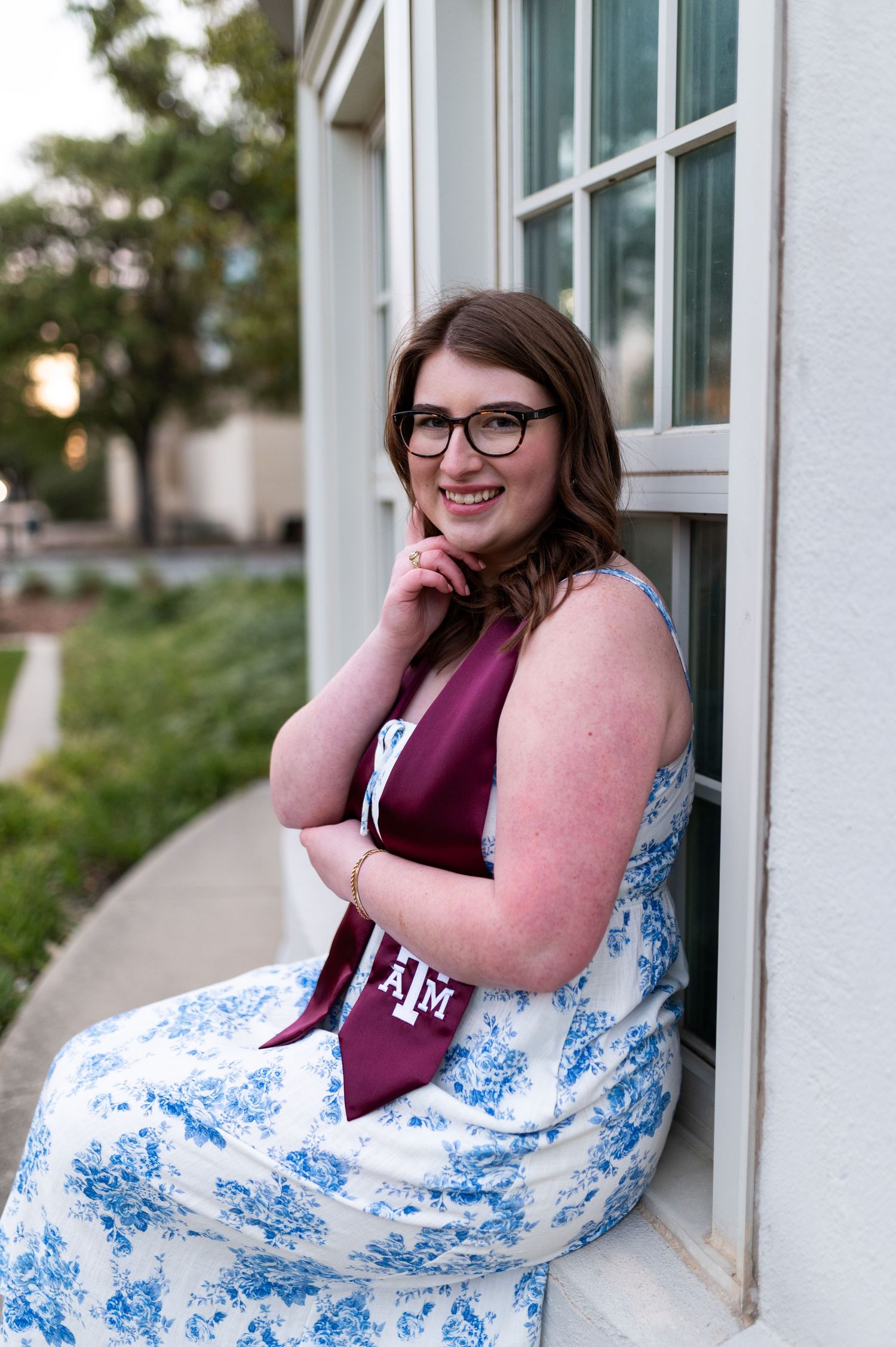 Texas A&M graduate smiling in floral dress with maroon ATM stole, seated by white window.