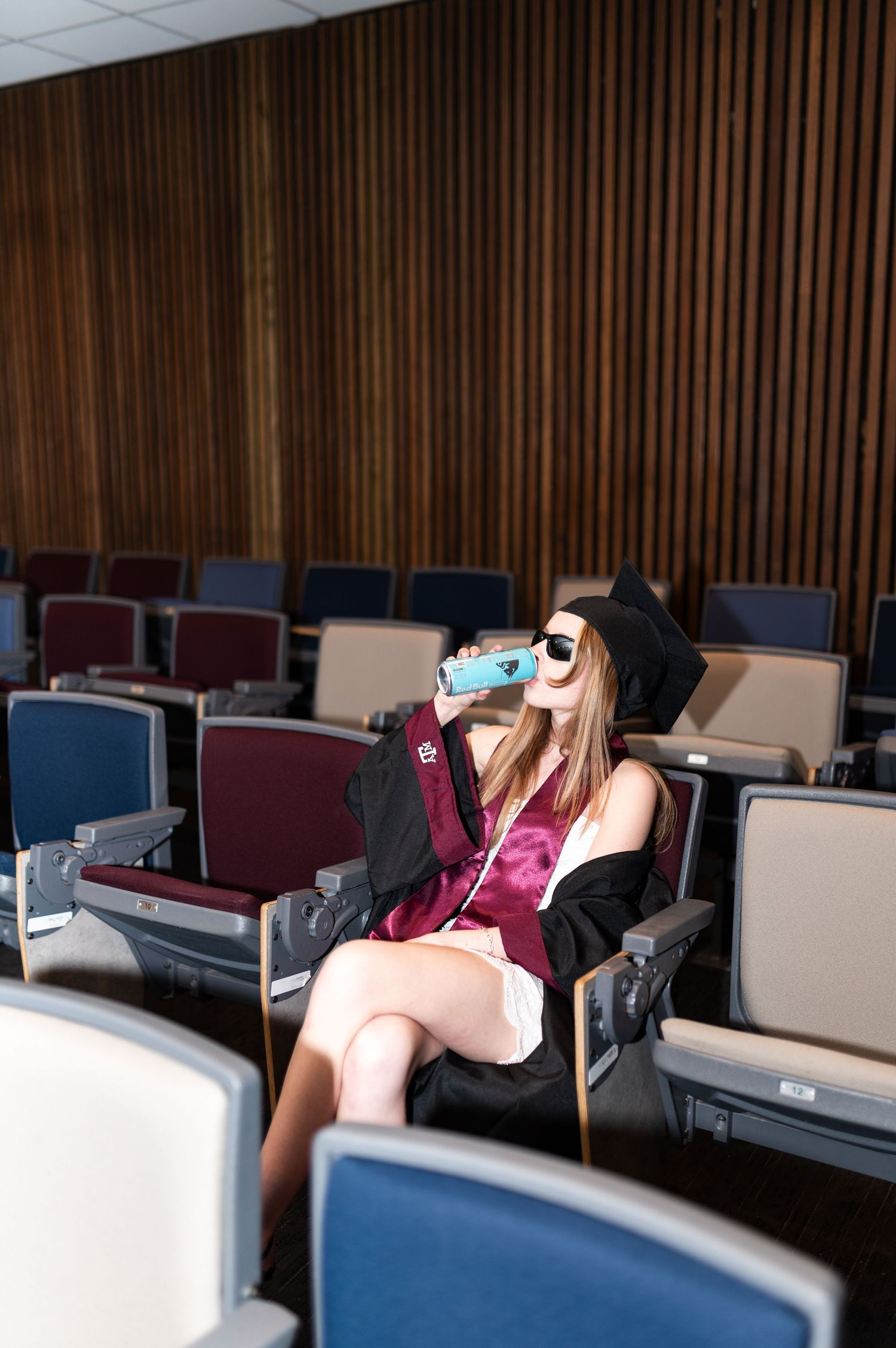 Texas A&M graduate in cap and gown sits in auditorium seats drinking from blue water bottle wearing sunglasses.