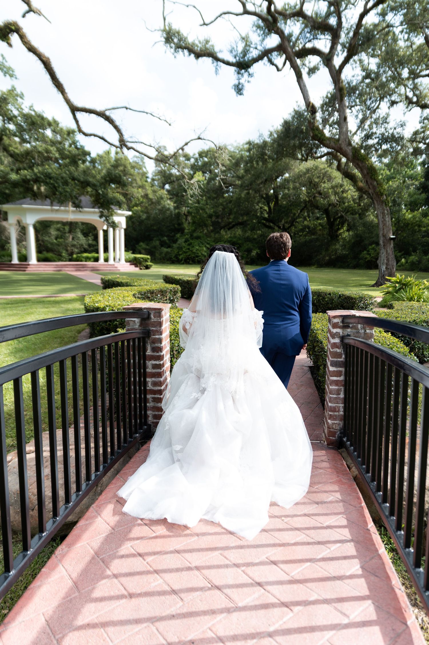 Bride and groom walking hand in hand across brick bridge toward garden gazebo at wedding venue.