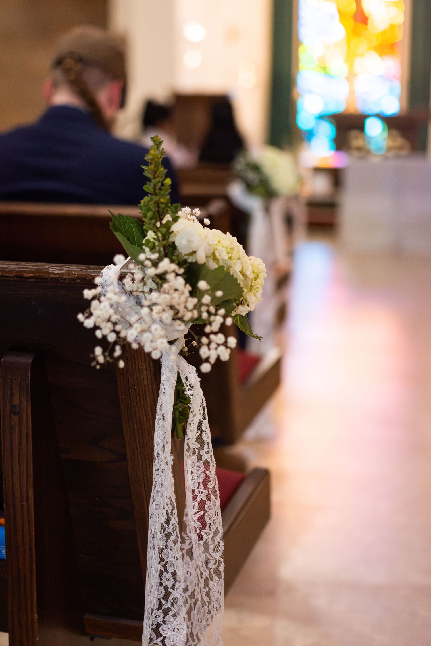White floral bouquet with baby's breath and lace ribbon decorating a wooden church pew for a wedding ceremony.