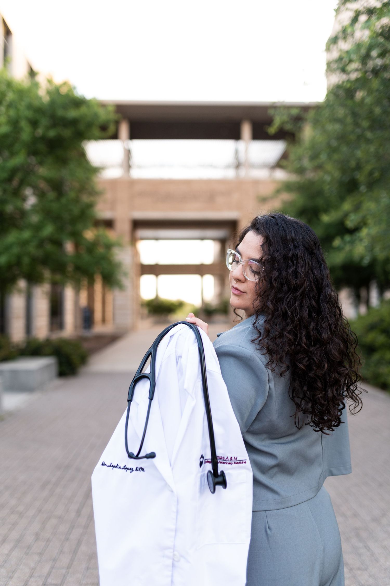 Female medical graduate carries white embroidered lab coat with stethoscope over shoulder outdoors.