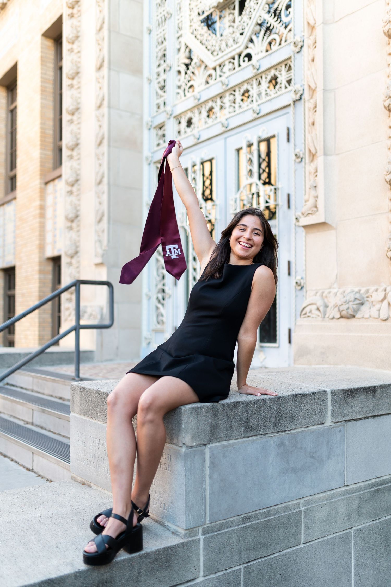 Happy graduate in black dress raising Texas A&M maroon stole while sitting on stone steps.