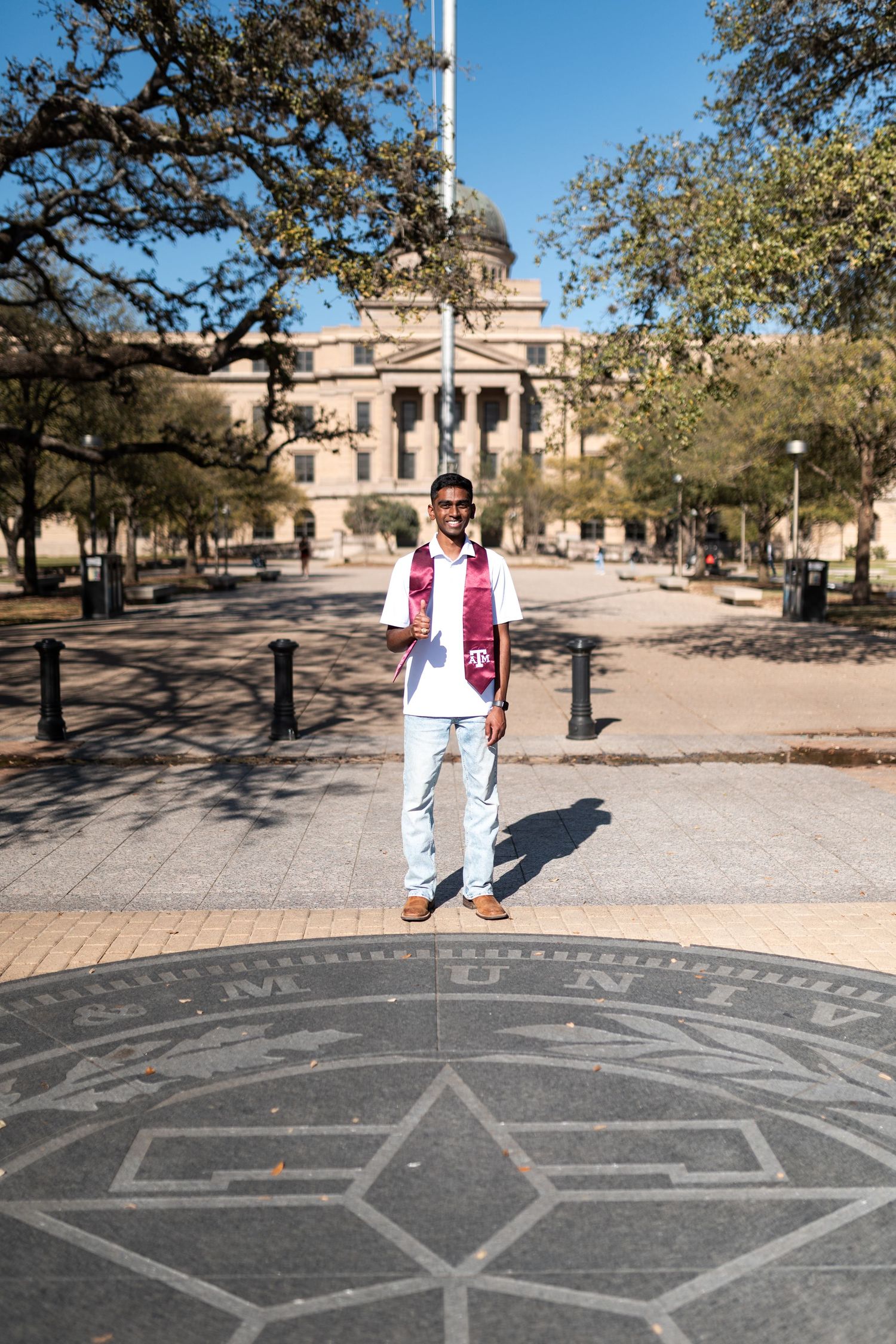 Texas A&M graduate stands on campus compass rose in front of historic Academic Building wearing maroon stole.