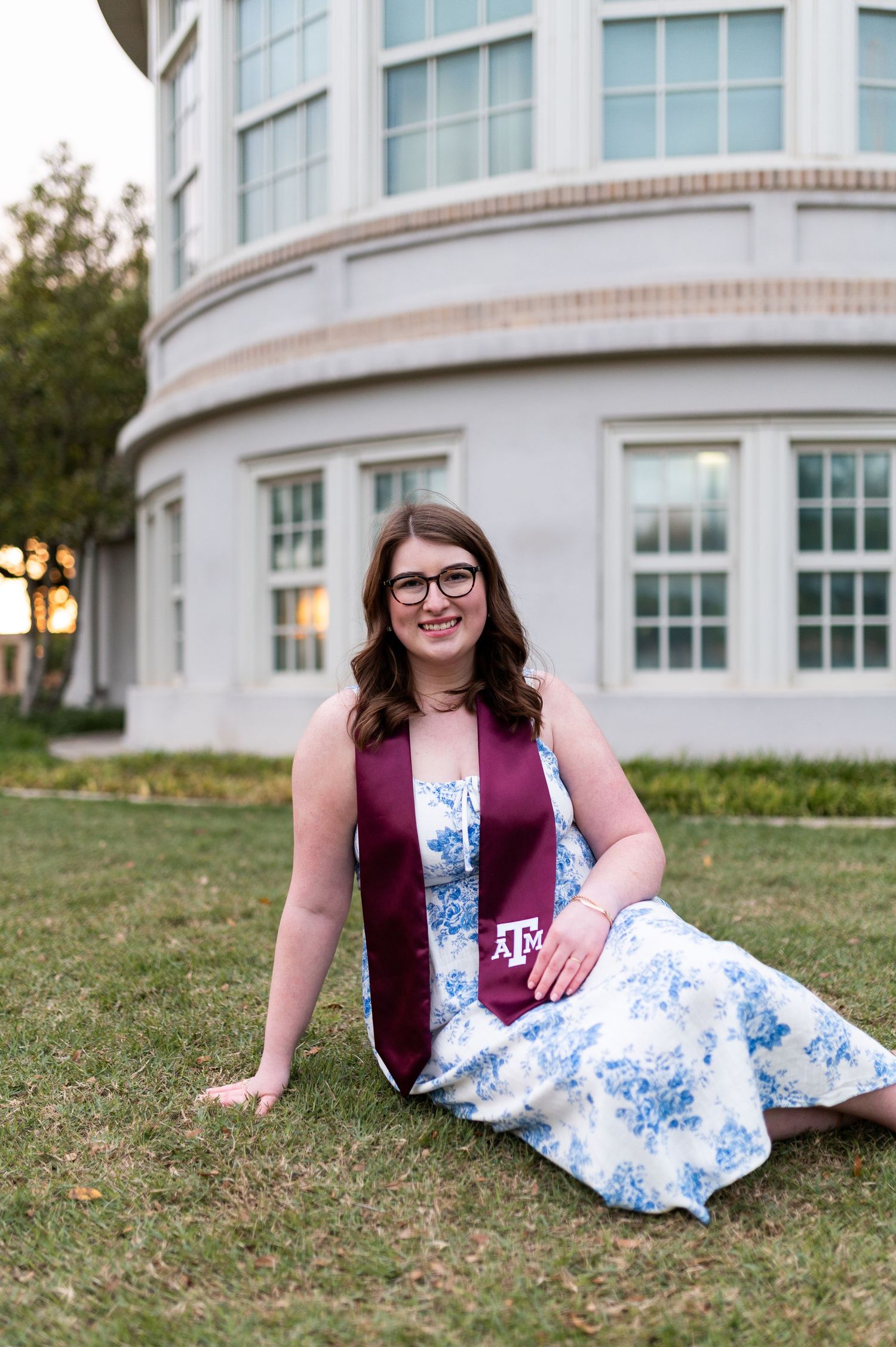 Smiling Texas A&M graduate in floral dress with maroon stole sitting on green campus lawn.