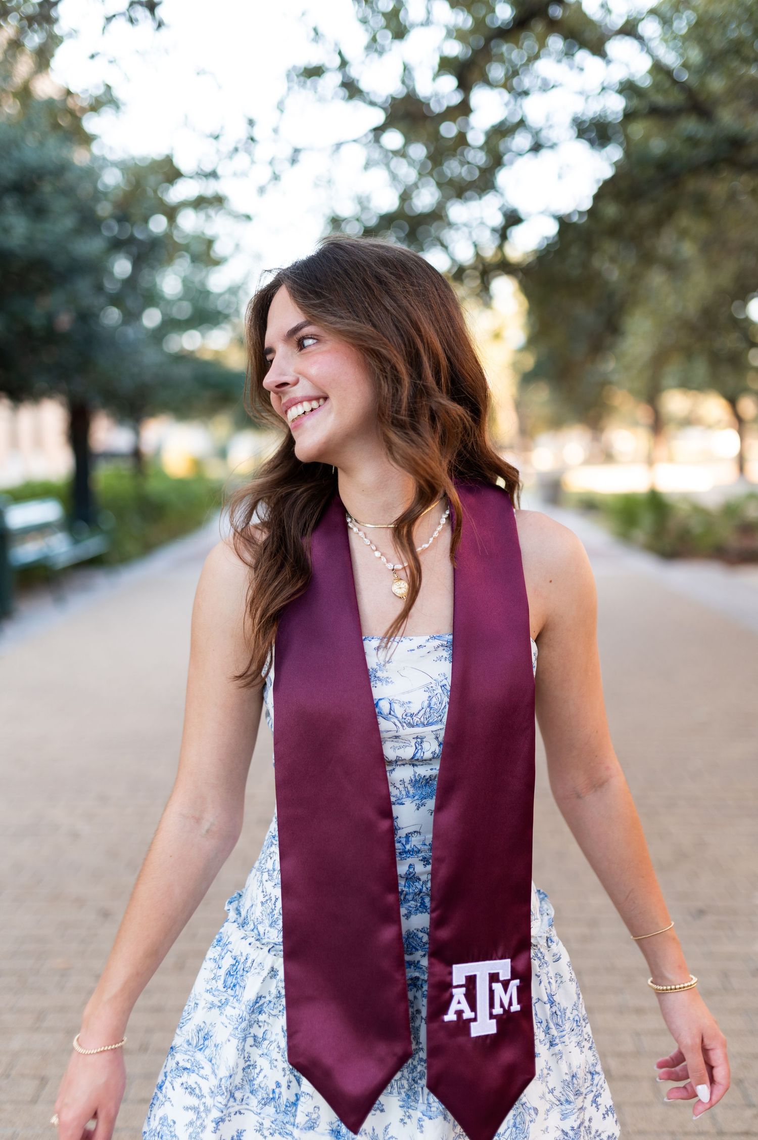 A smiling young woman wears a maroon Texas A&M graduation stole over a floral dress on a tree-lined campus path.