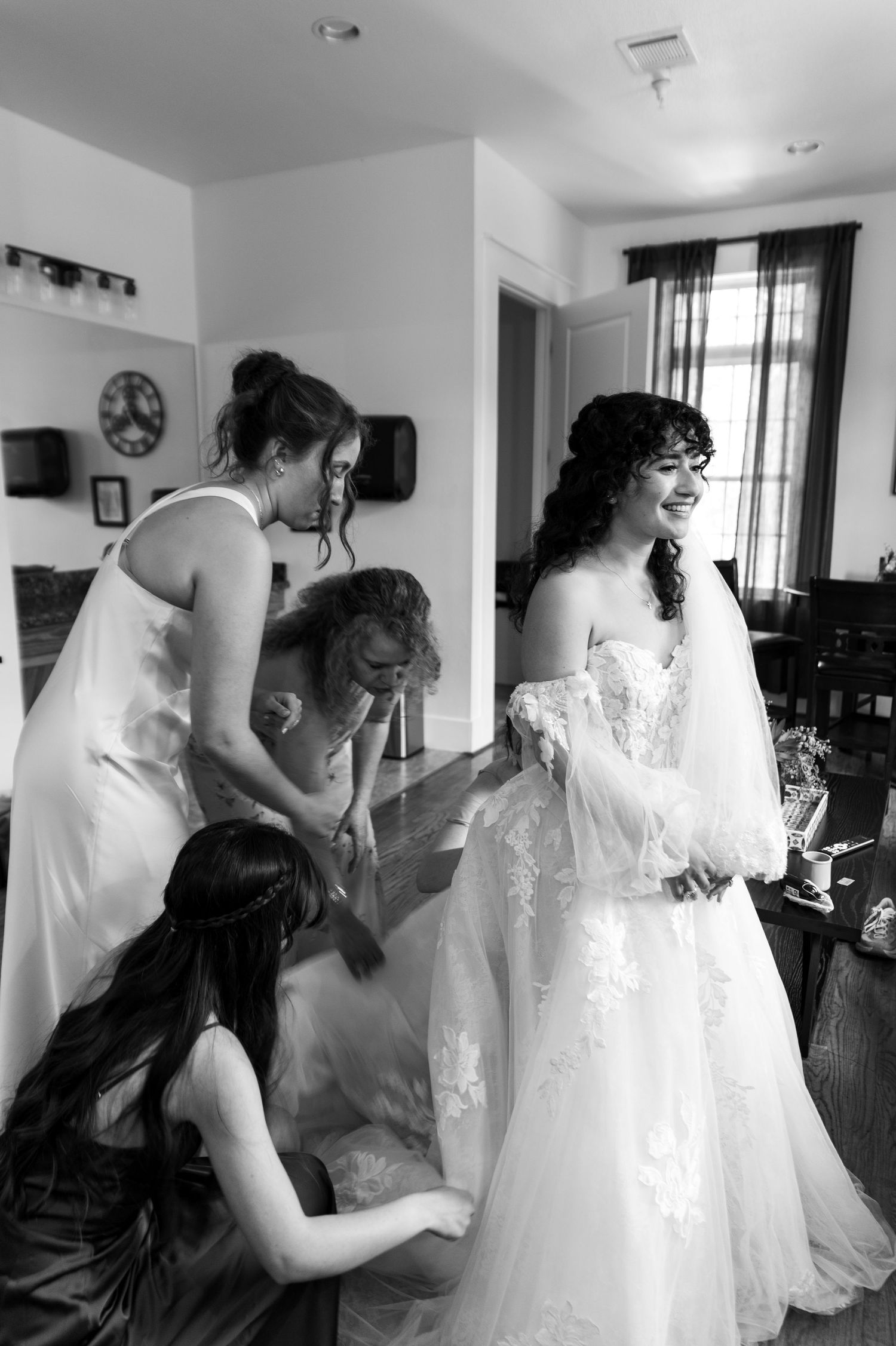 Bridesmaids help smiling bride into lace wedding gown during bridal preparations in black and white photo.