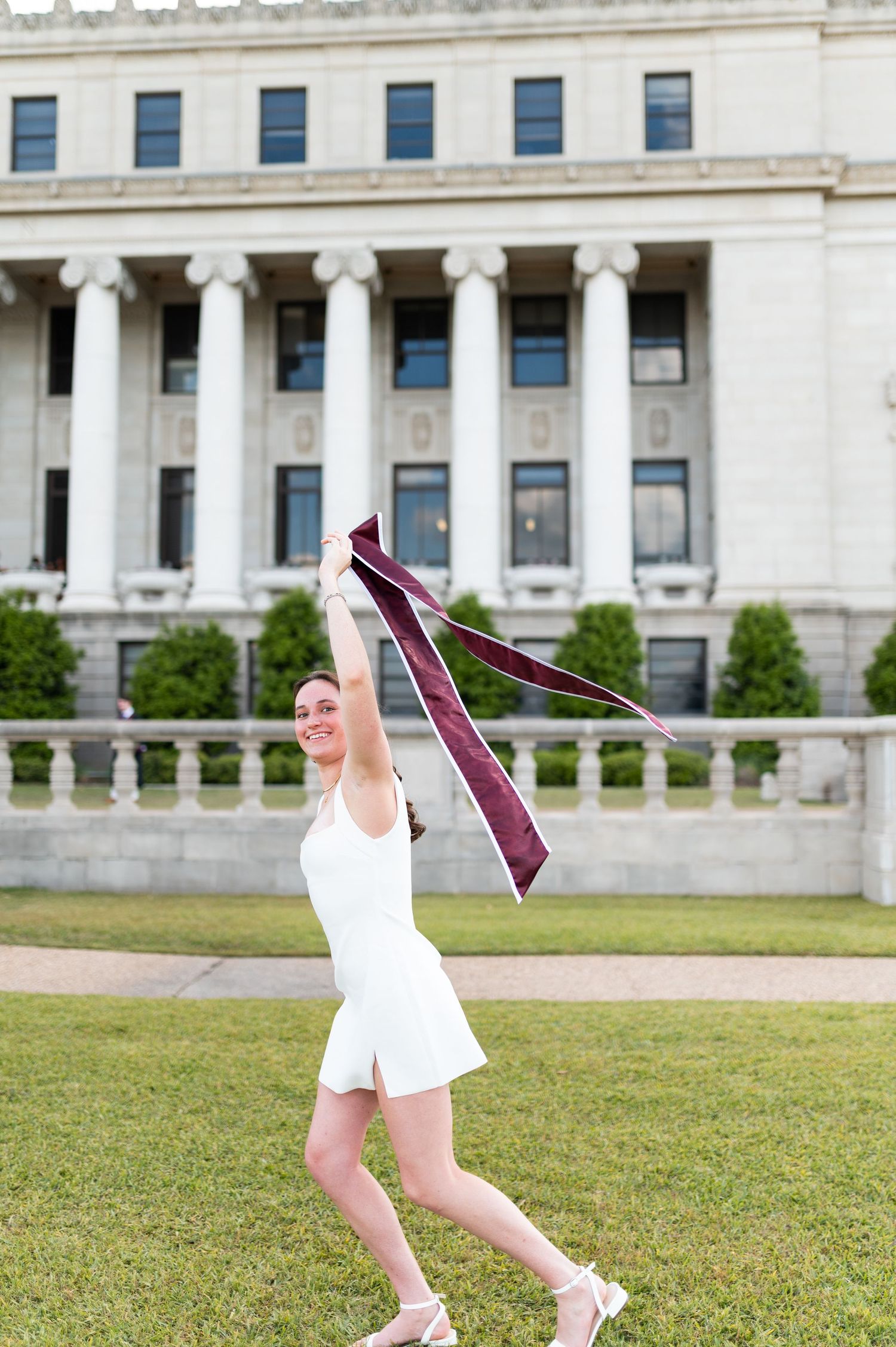 Joyful Texas A&M graduate waves maroon stole in air while standing on campus lawn in white dress. Grad Photos at the Admin building