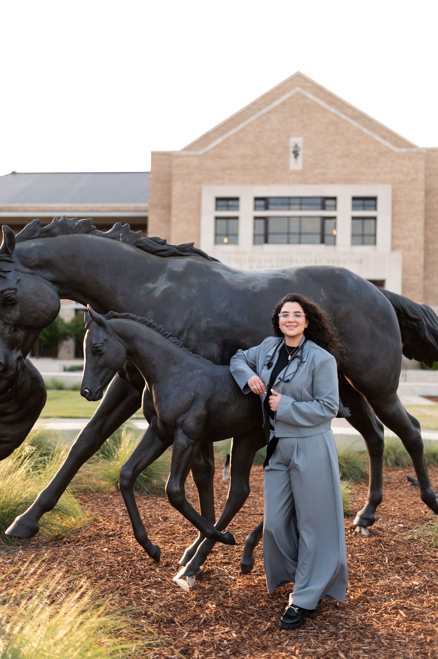 Smiling woman in gray pantsuit poses beside bronze running horse sculptures at Texas university campus.