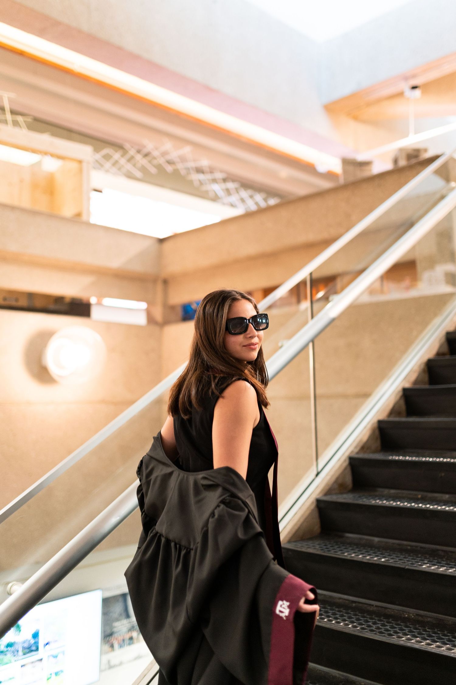 Young woman in black dress and sunglasses carrying graduation stole on indoor escalator.