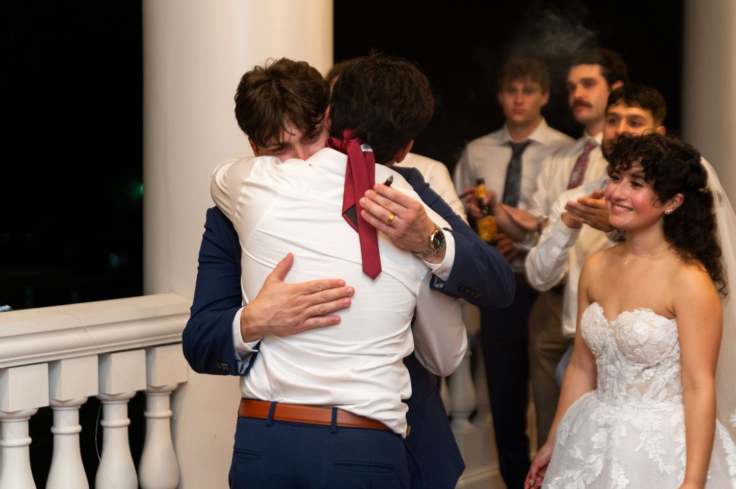 Groom in navy suit embracing guest while bride in lace gown watches at outdoor wedding reception.