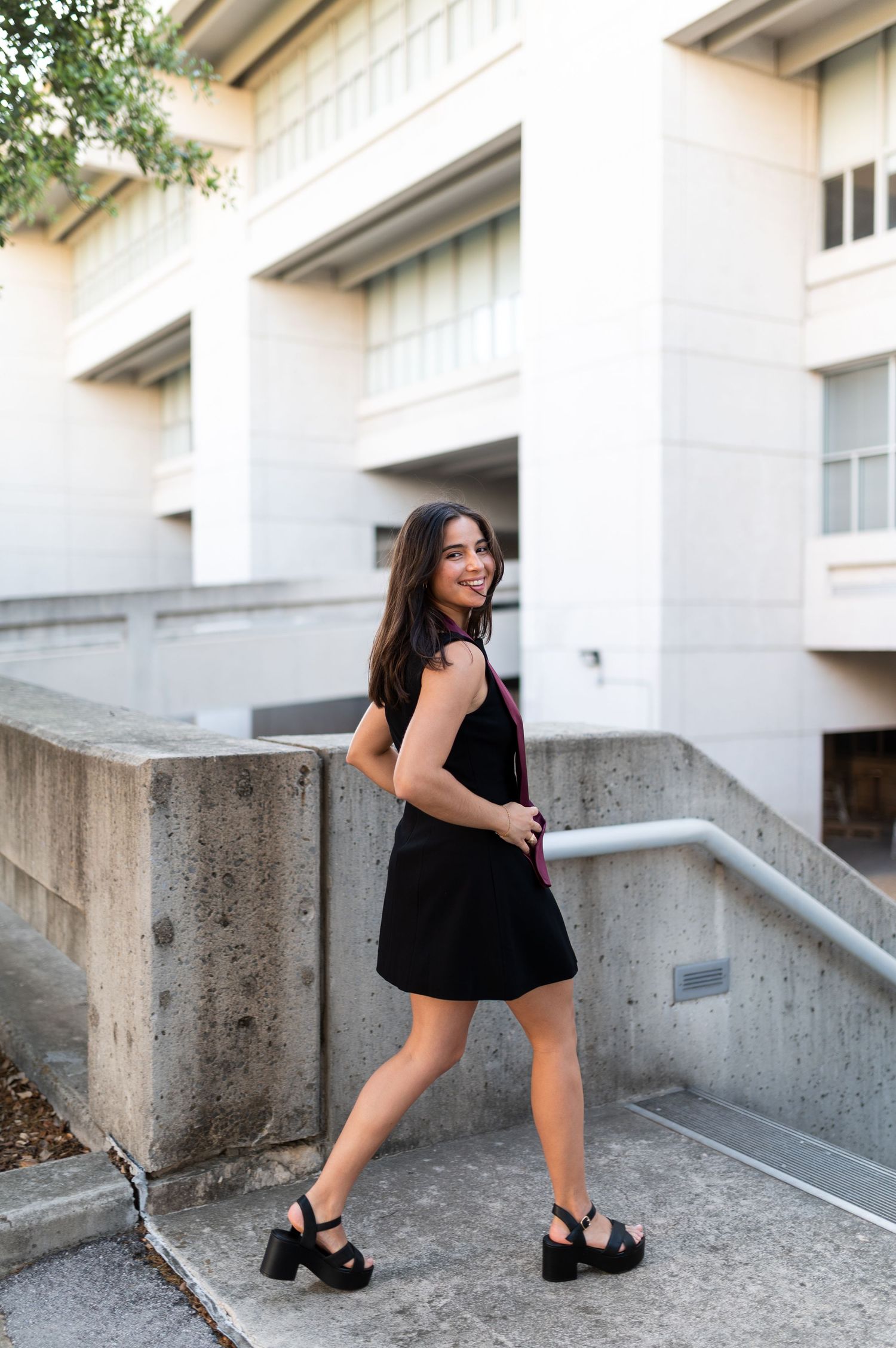 Young woman in black mini dress and platform sandals poses on outdoor campus stairs smiling over shoulder.