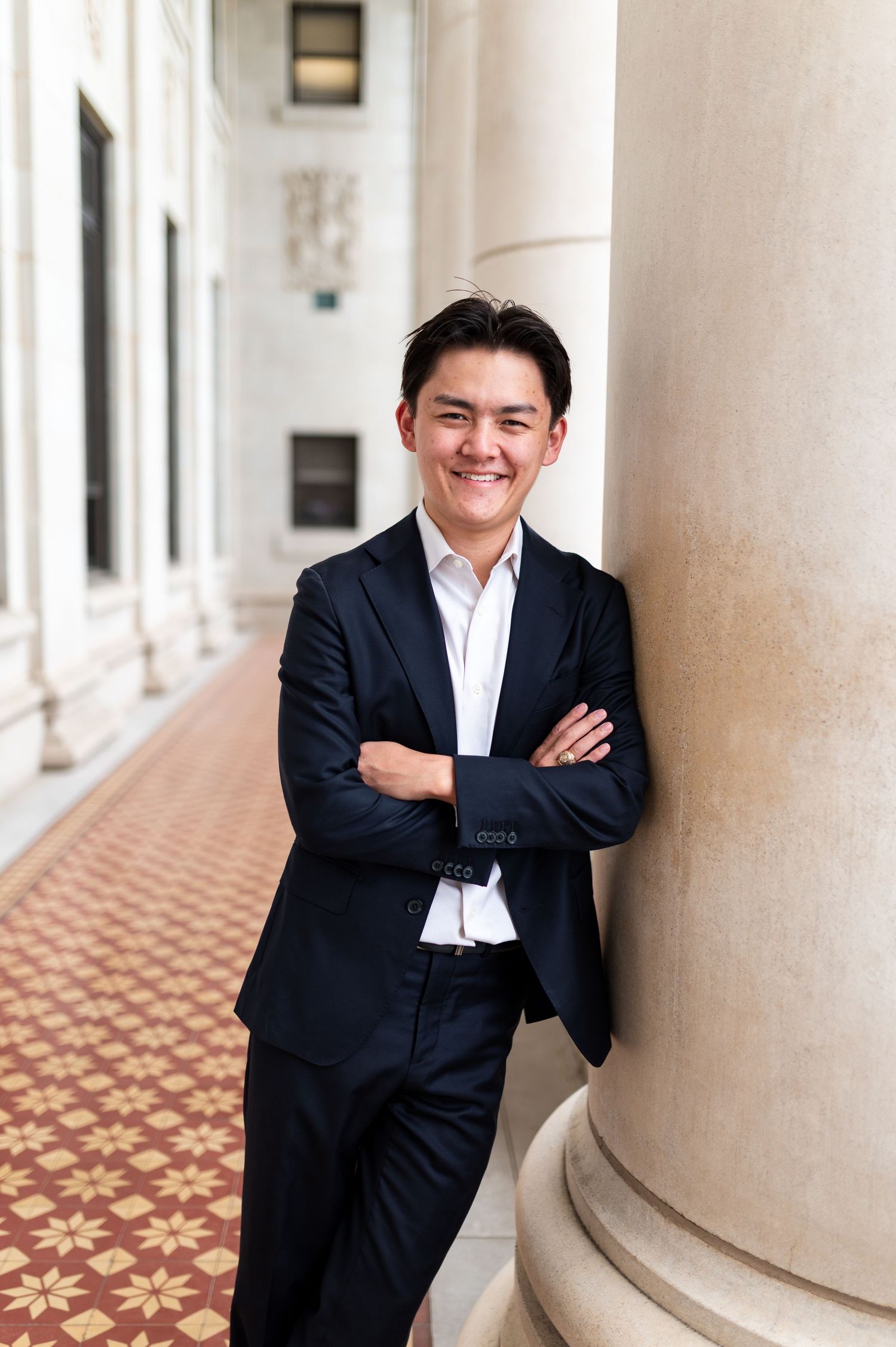 Smiling young man in navy suit leaning against column in elegant columned hallway. Grad Photos at the Texas A&M Admin building