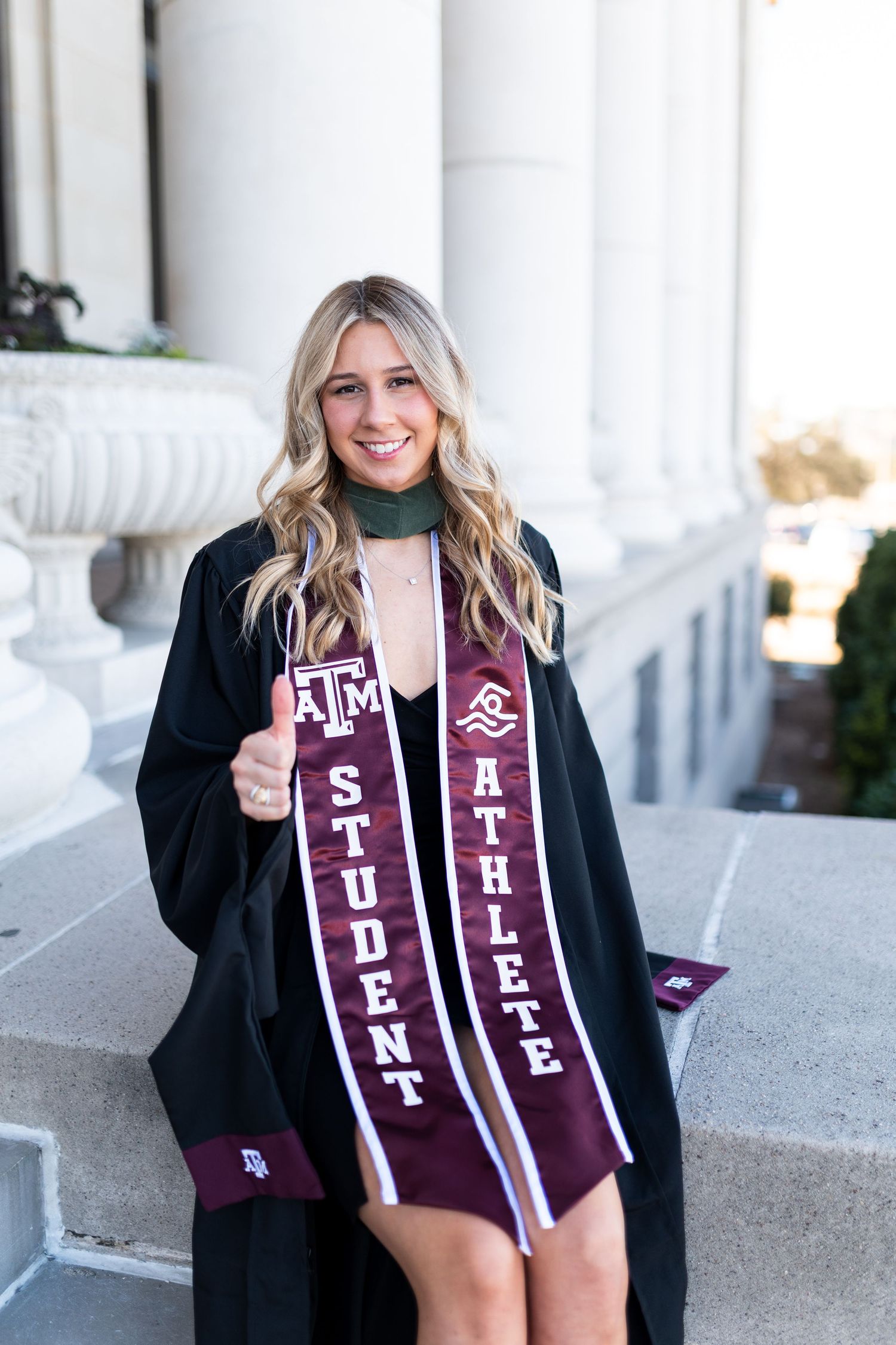 Texas A&M student athlete smiles in graduation gown with maroon Student Athlete sash, giving thumbs up. Grad Photos at the Admin building