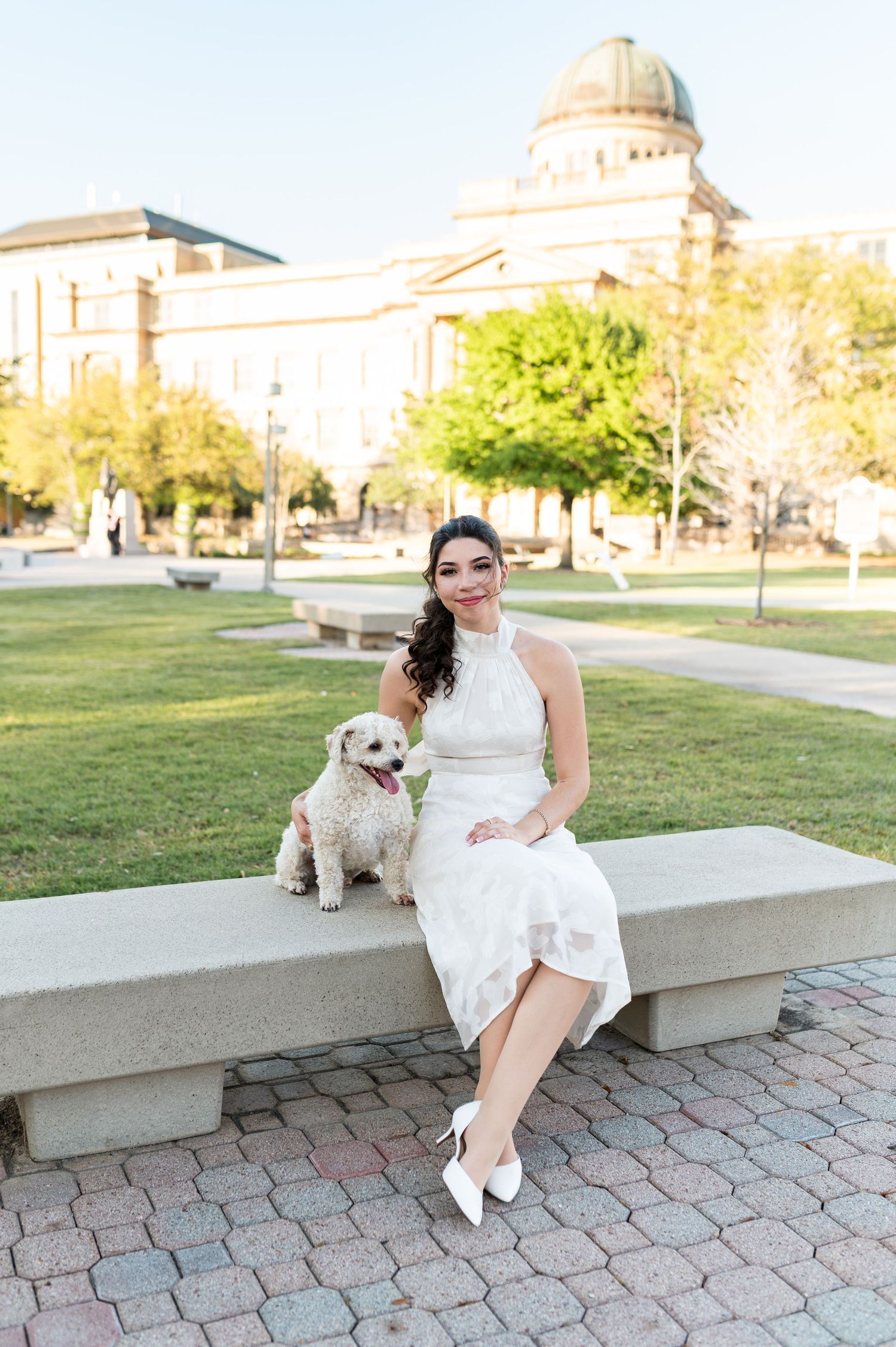 Smiling bride in white dress sits on stone bench with fluffy white dog, Academic Building in background.