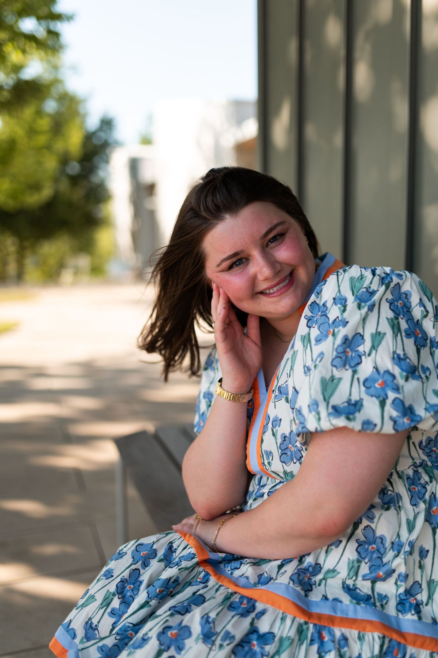 Young woman in blue floral dress with orange trim smiling while seated outdoors near pillars.