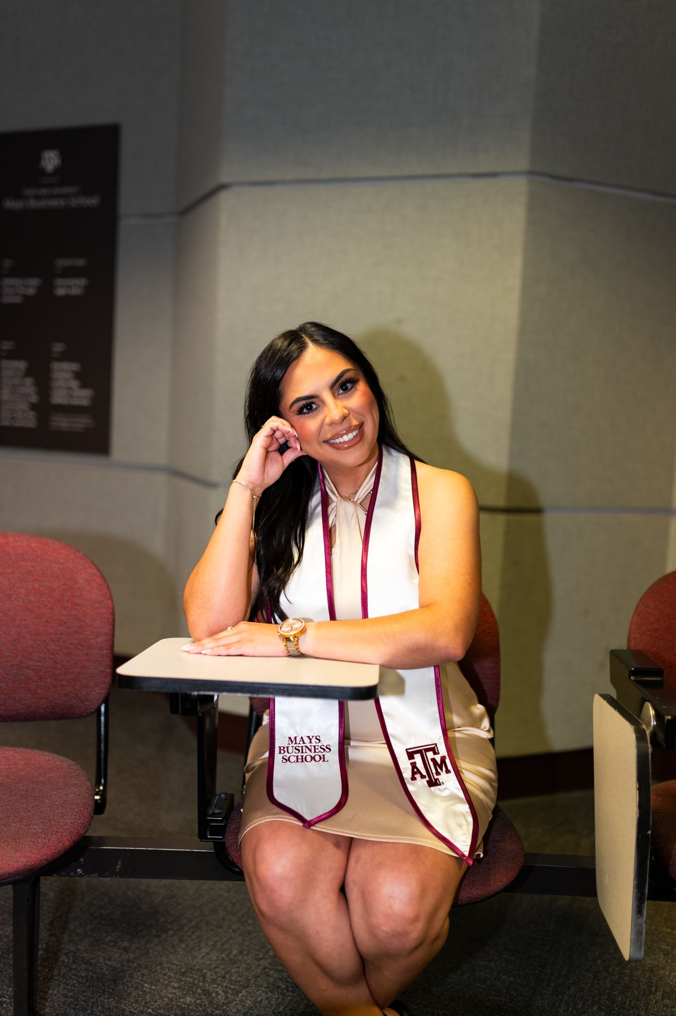 Female Texas A&M Mays Business School graduate poses at desk wearing white graduation stole indoors.