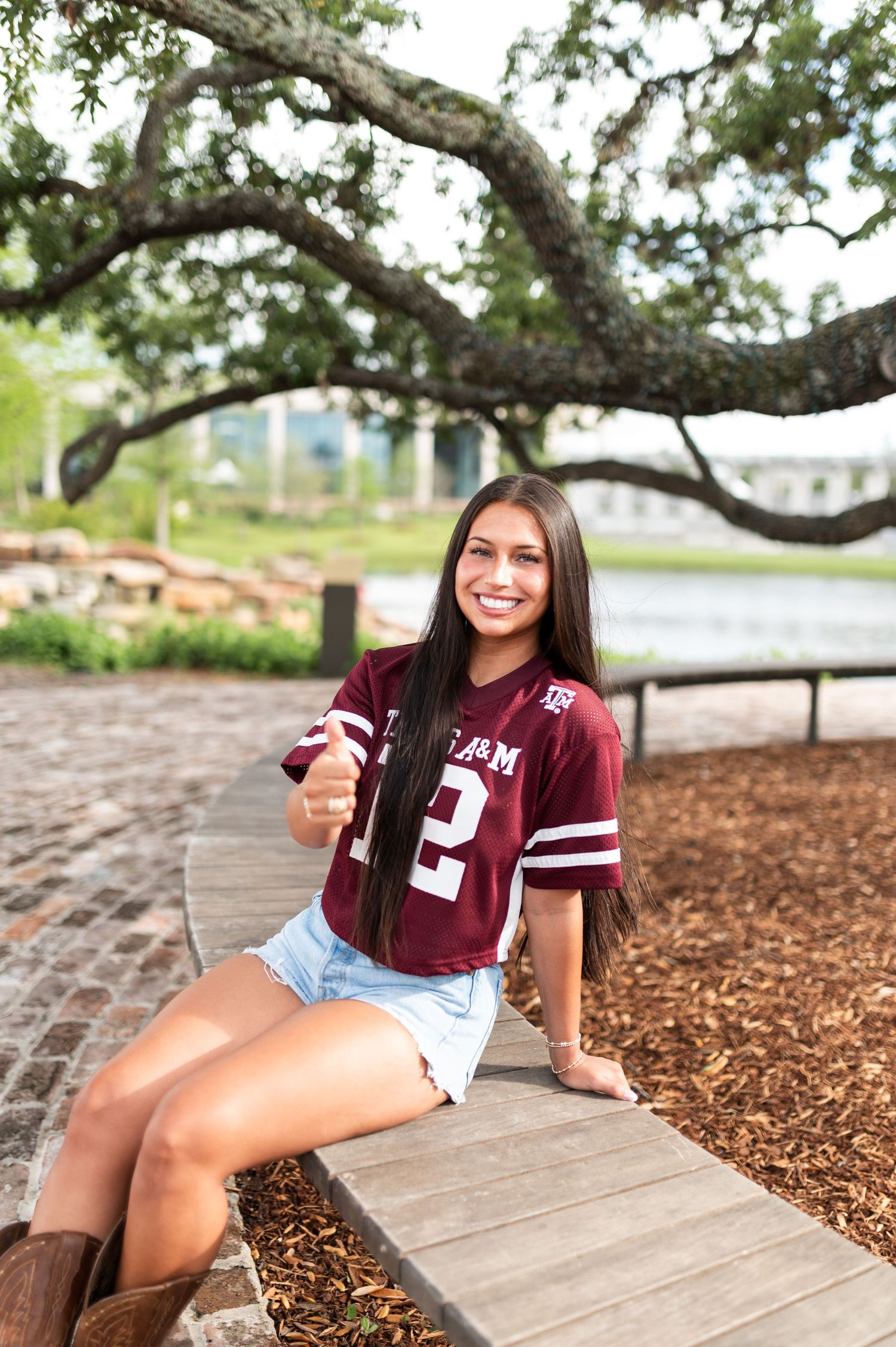 A smiling young woman in a Texas A&M jersey sitting on a bench under a large oak tree.
