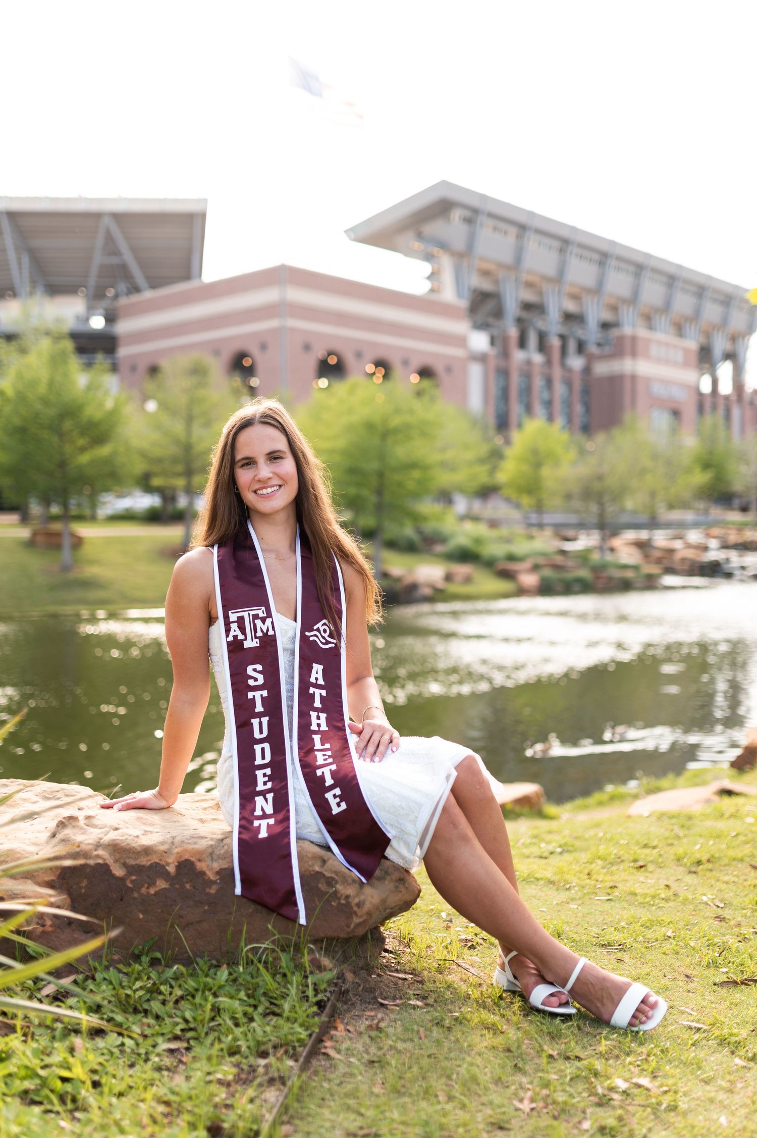 Texas A&M female graduate in white dress sits on rock near campus pond wearing Student Athlete sash.