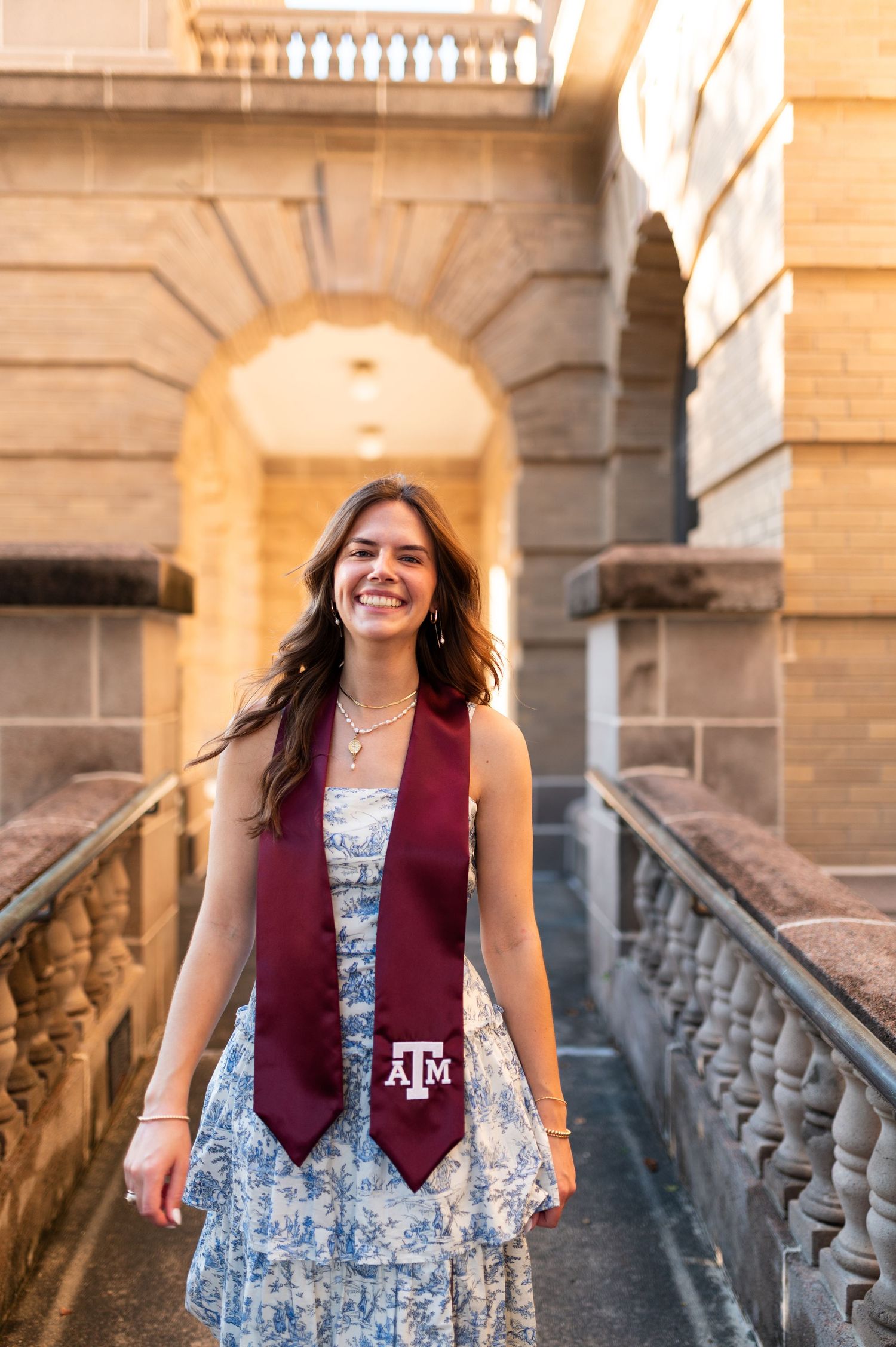 Texas A&M graduate smiles in golden hour light beneath stone archway wearing maroon graduation stole.