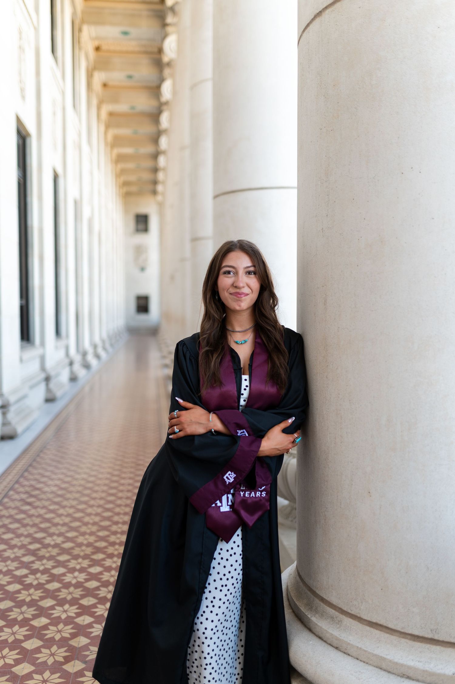 Woman in graduation gown and Texas A&M stole leans against column in colonnade. Grad Photos at the Admin building