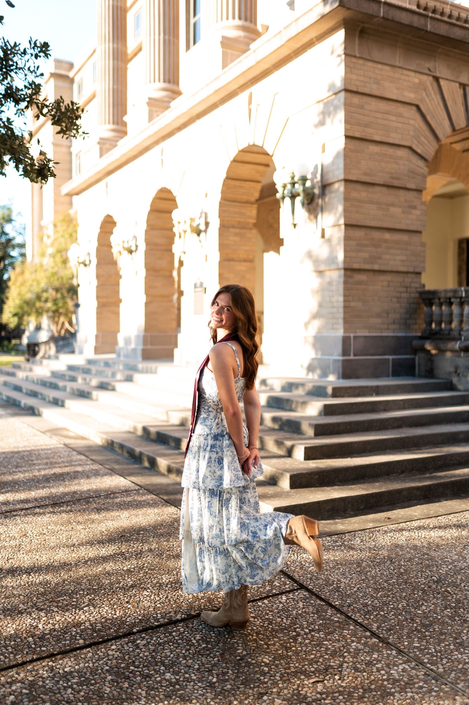 Young woman in floral maxi dress and cowboy boots twirls outside historic Texas A&M campus building.