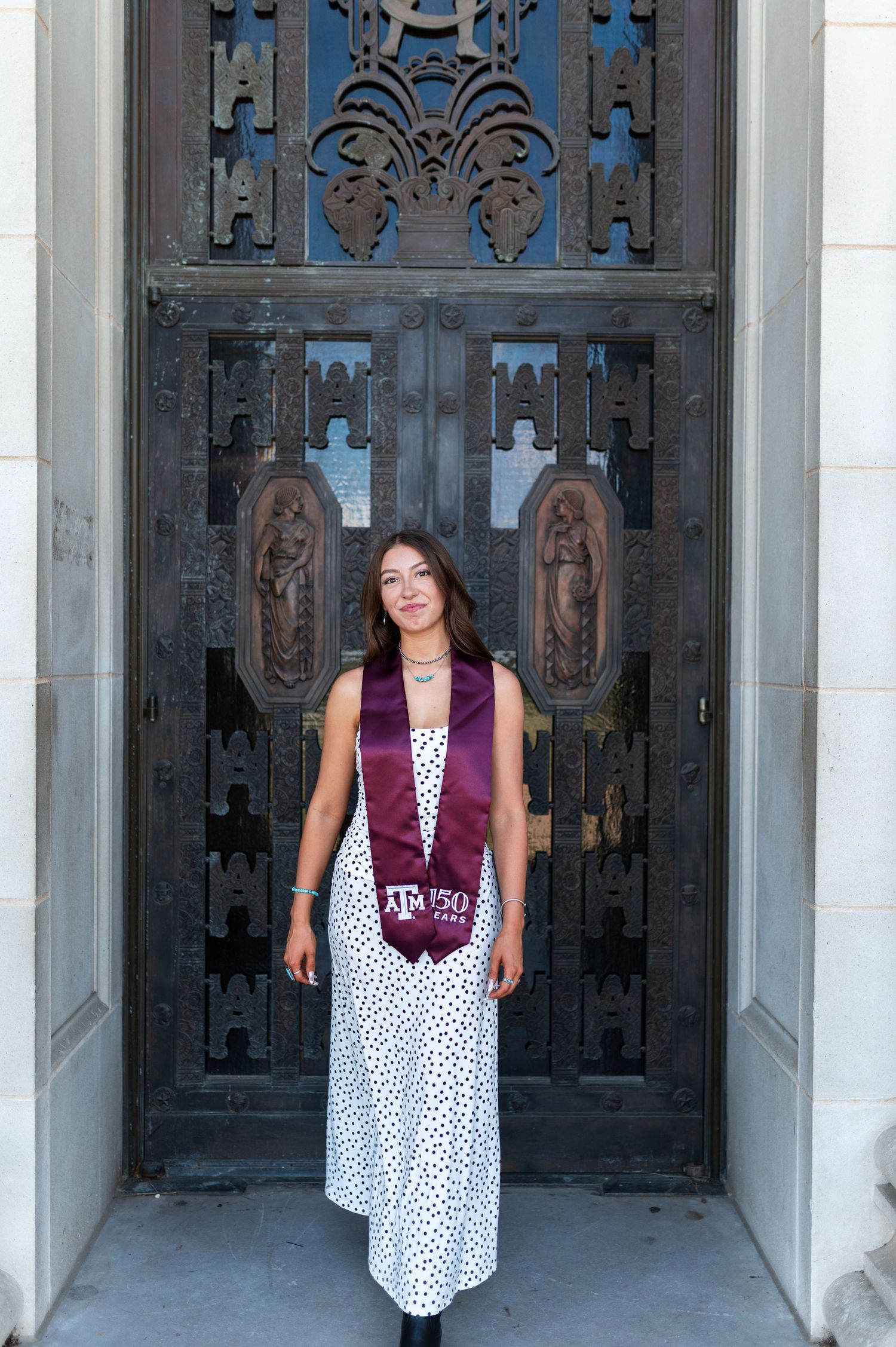 Texas A&M graduate in white polka dot dress and maroon 150 Years stole poses before ornate bronze doors. Grad Photos at the Admin building