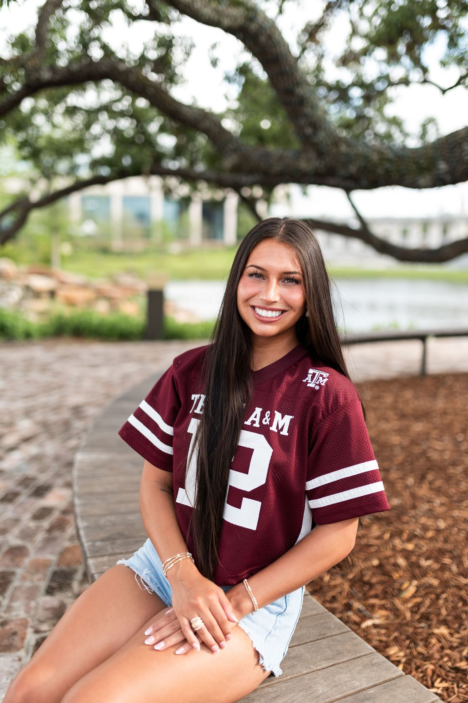 A smiling young woman in a Texas A&M jersey sitting on a wooden walkway outdoors.