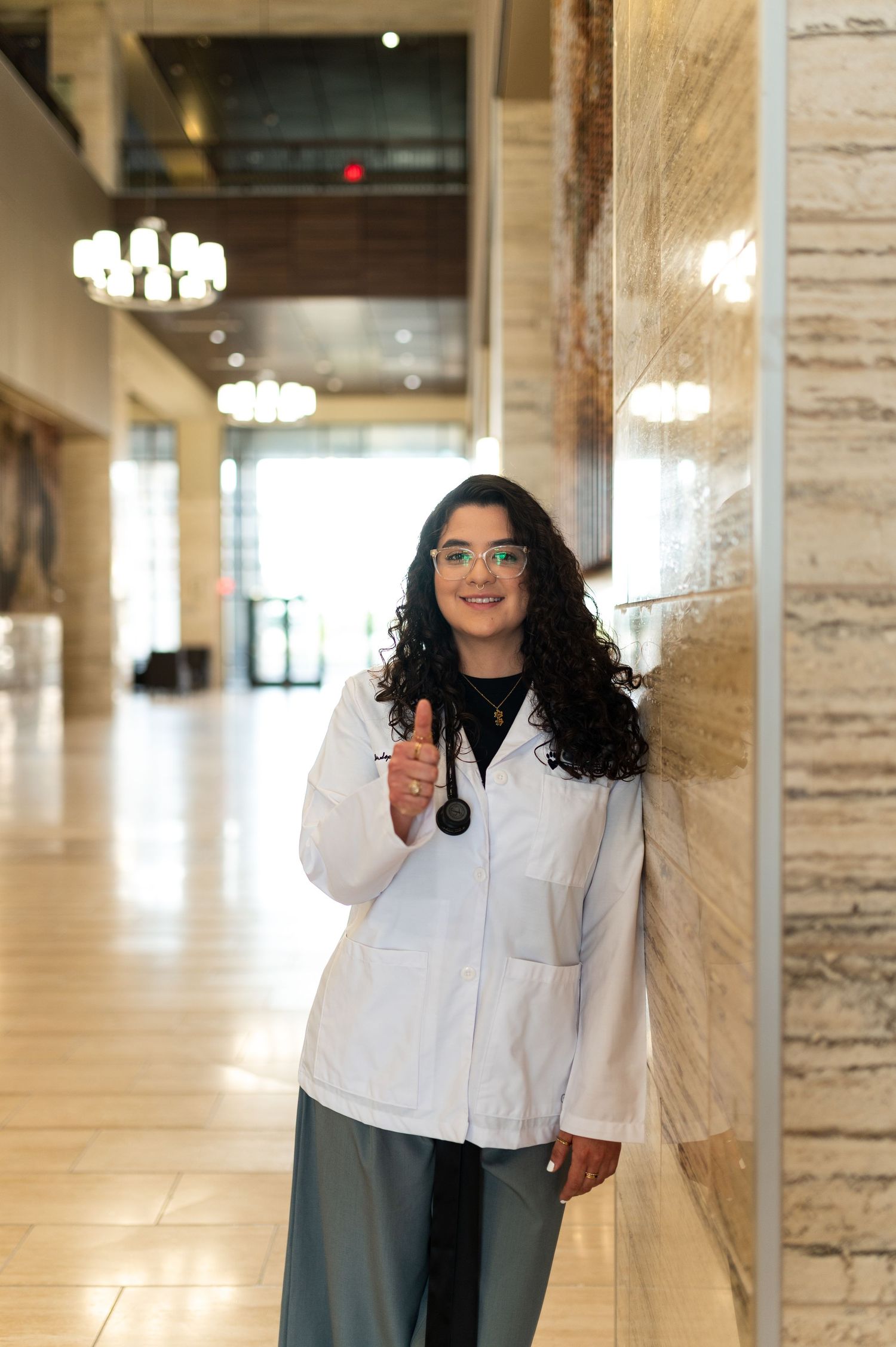 Female medical student in white lab coat with stethoscope poses in elegant marble hallway interior.