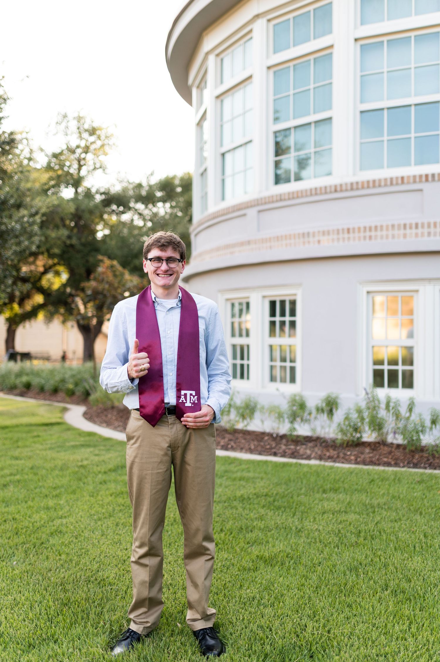 Smiling young man in khakis displays Texas A&M graduation stole on green lawn.