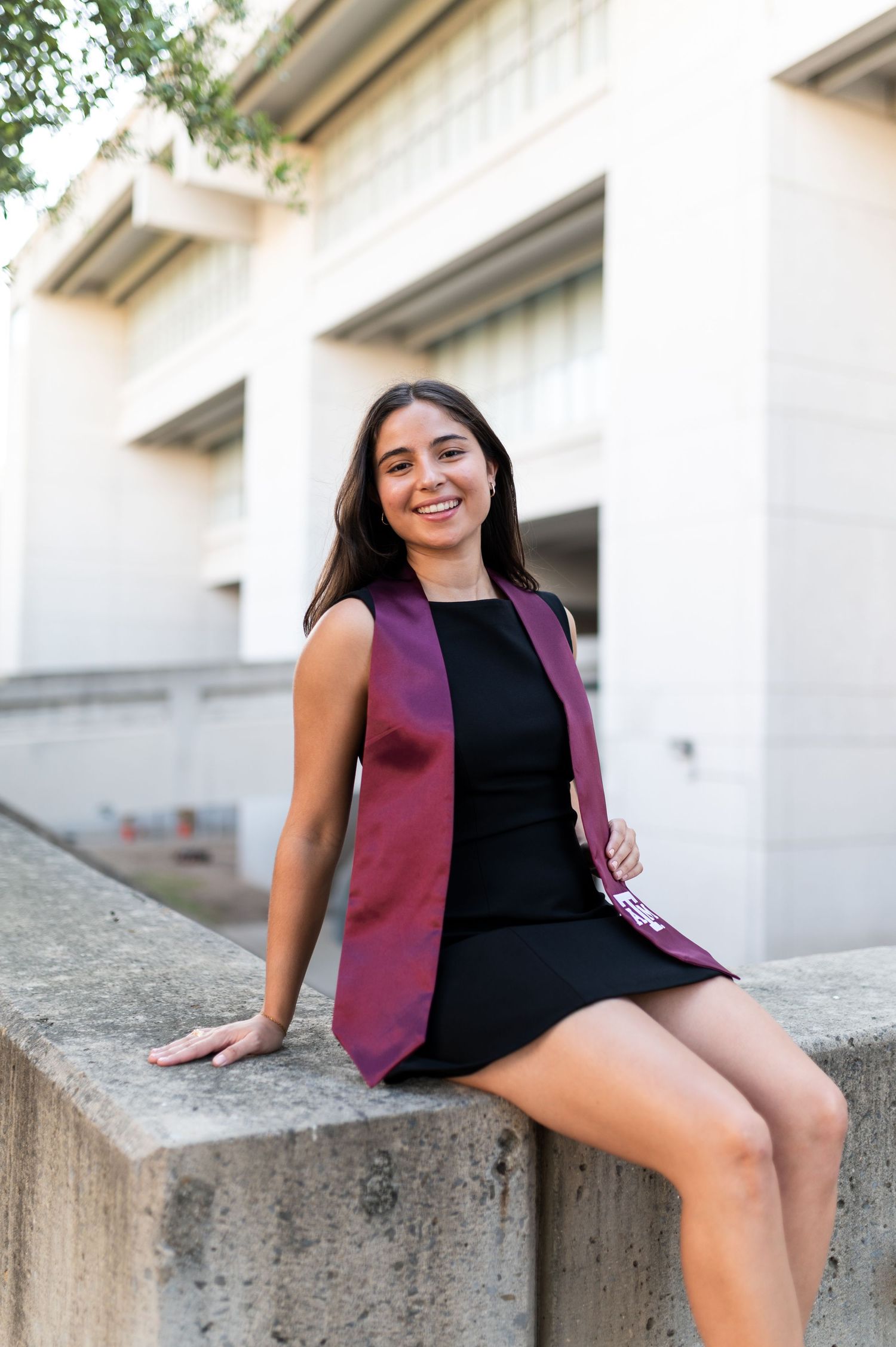 Young woman in black dress and maroon graduation stole sitting on concrete ledge outdoors smiling.