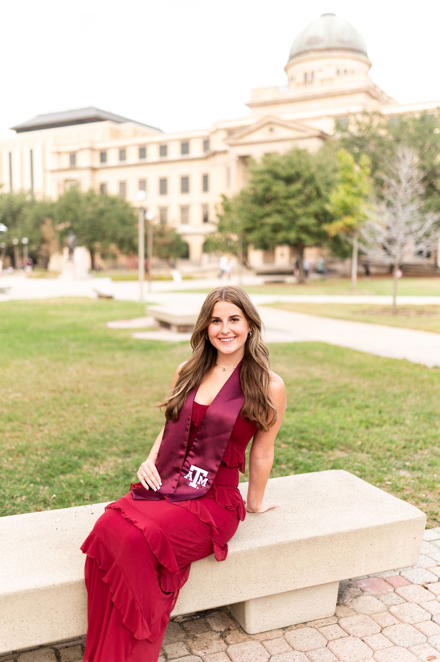 Smiling woman in red dress and Texas A&M maroon stole sits on stone bench with Academic Building behind her.