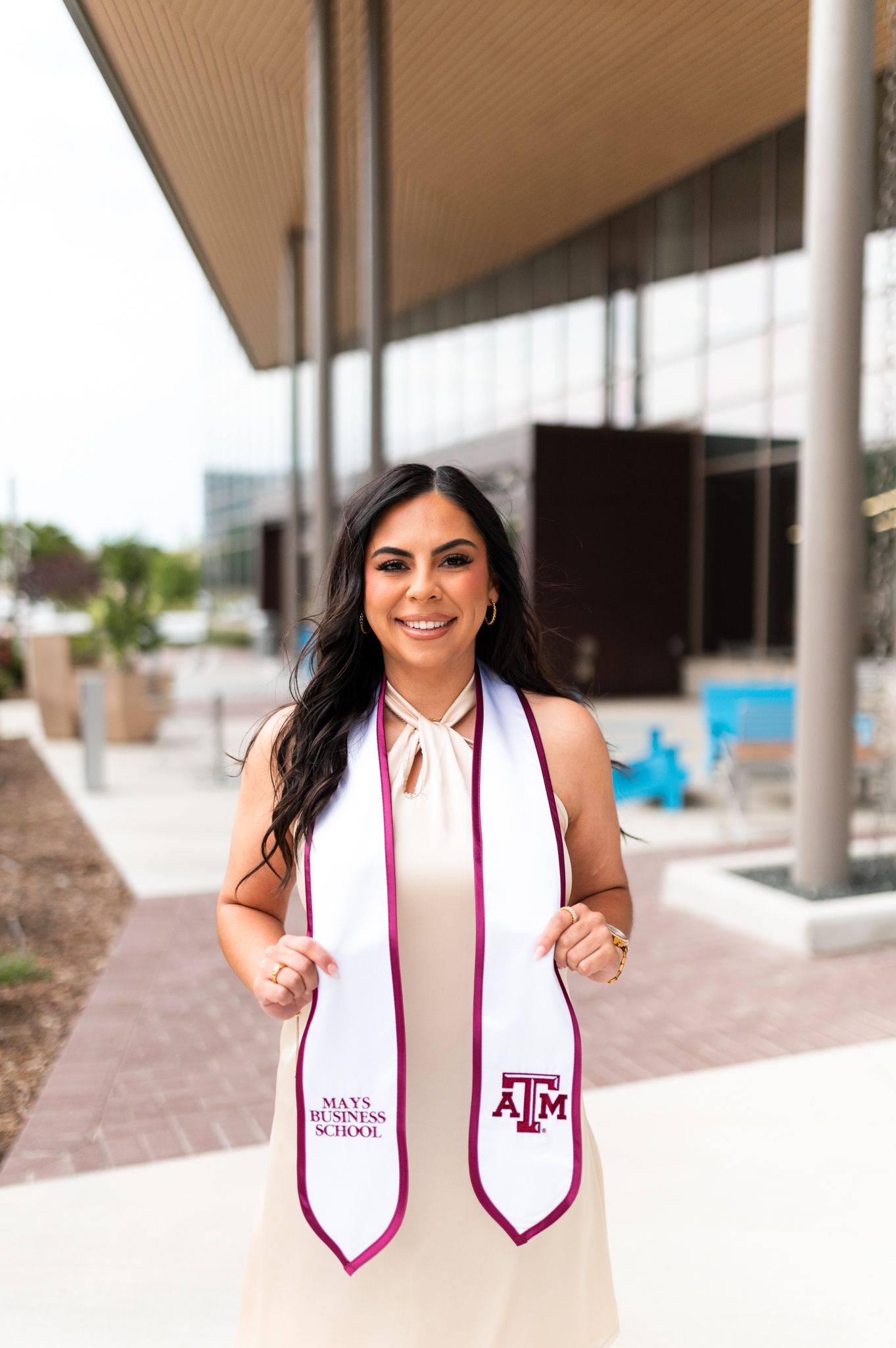 Smiling female Texas A&M Mays Business School graduate holds white maroon-trimmed stole outdoors.