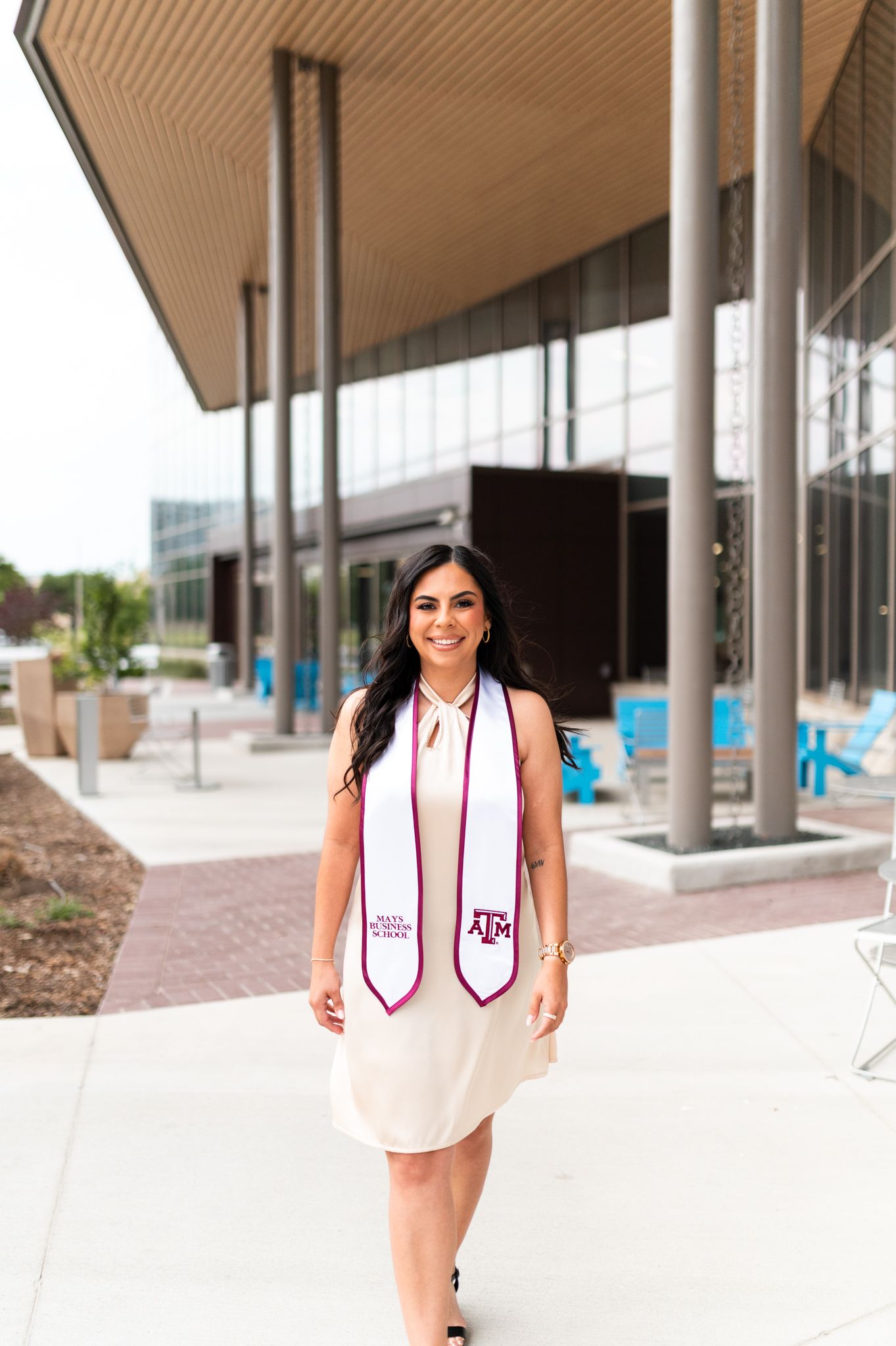 Smiling woman in white dress with Texas A&M graduation stole outside modern university building.