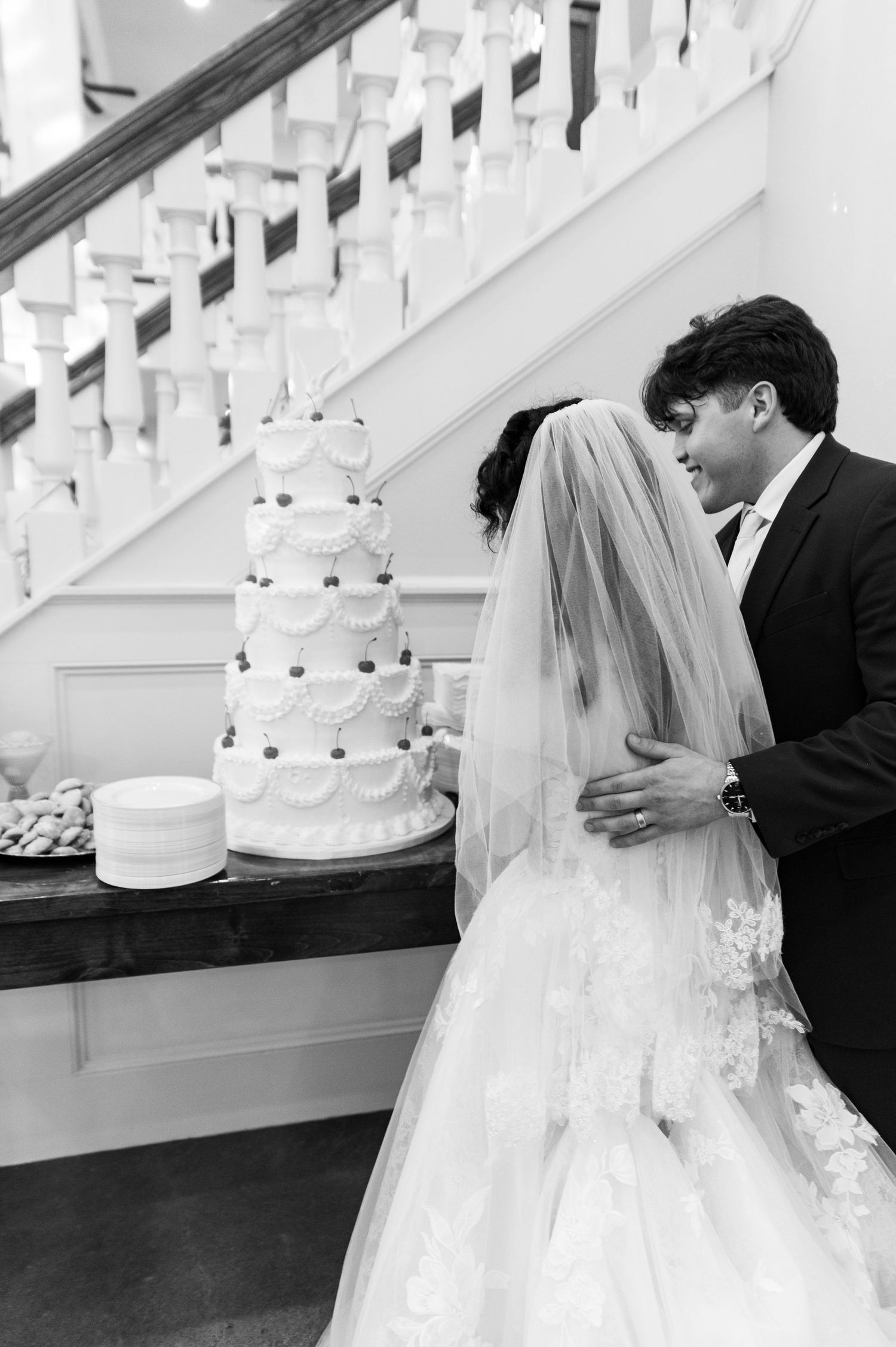 Black and white photo of bride and groom sharing intimate moment beside tiered wedding cake.