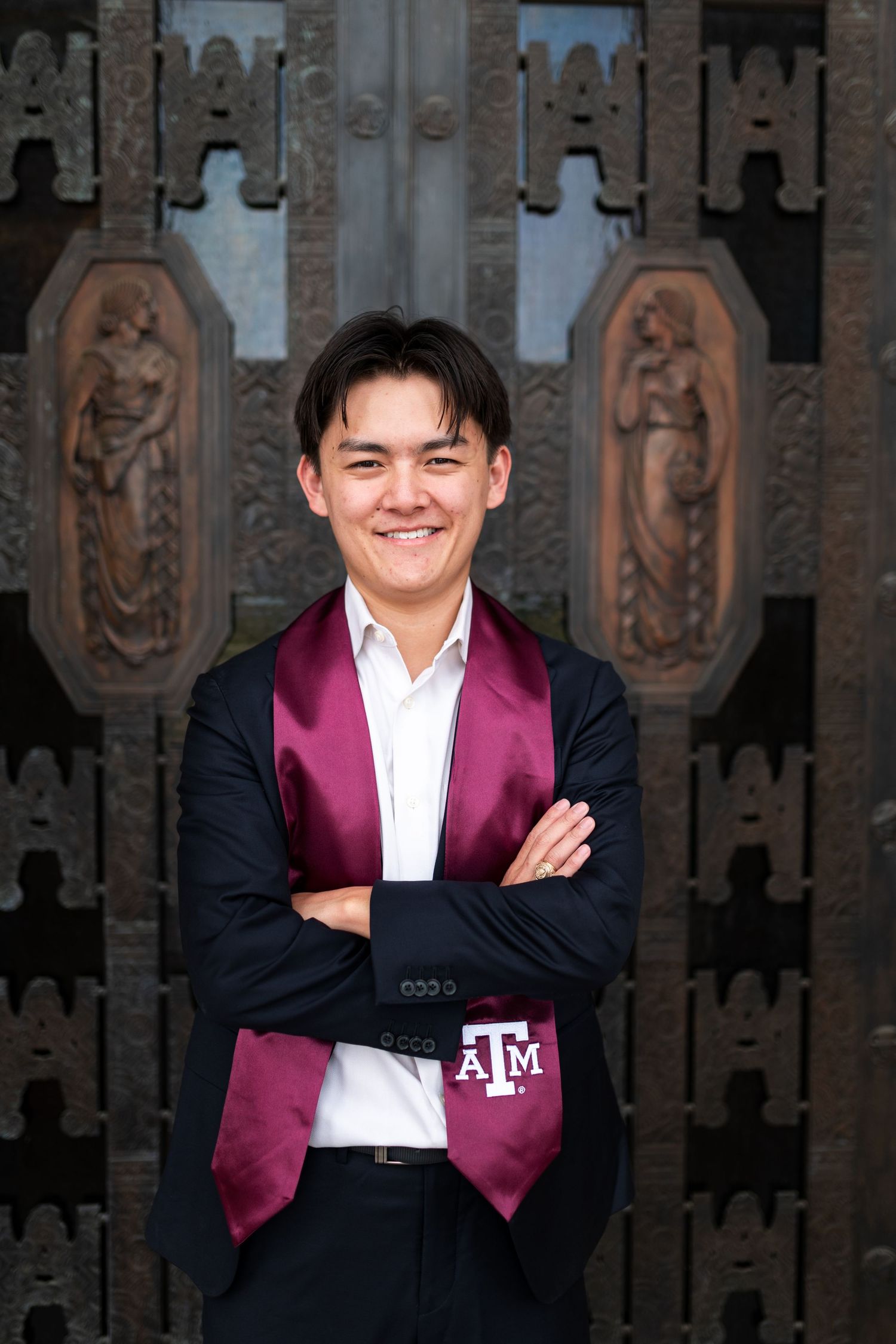 Texas A&M graduate in navy suit with maroon stole poses arms crossed before ornate campus doors. Grad Photos at the Admin building