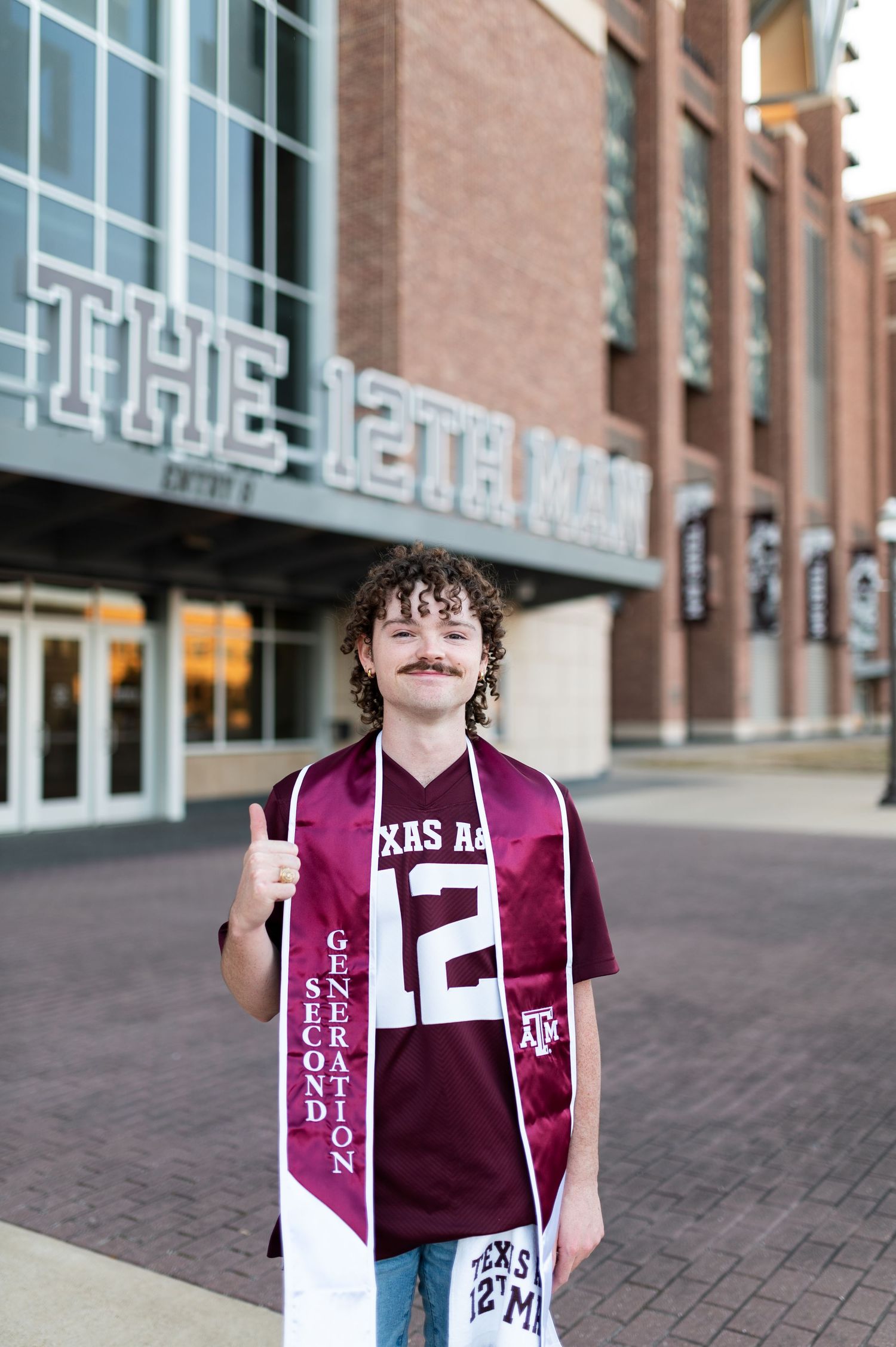 Texas A&M graduate in maroon jersey and Second Generation stole gives thumbs up outside The 12th Man stadium.