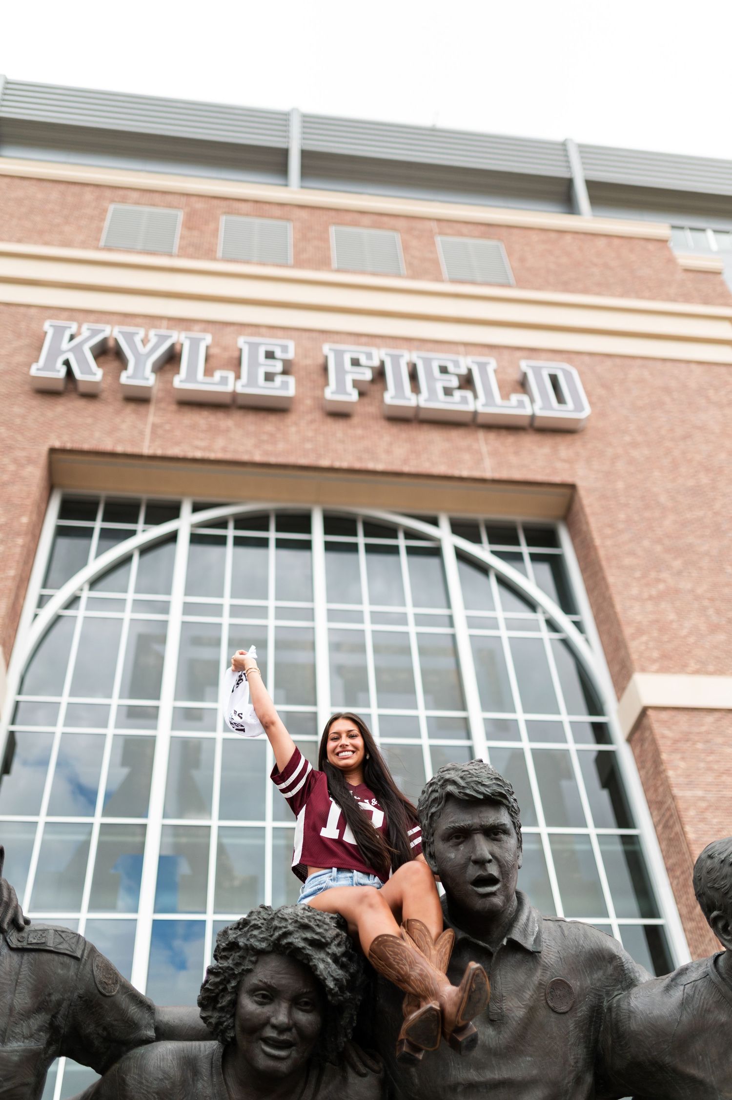 Texas A&M fan in maroon jersey poses on bronze statues in front of Kyle Field.