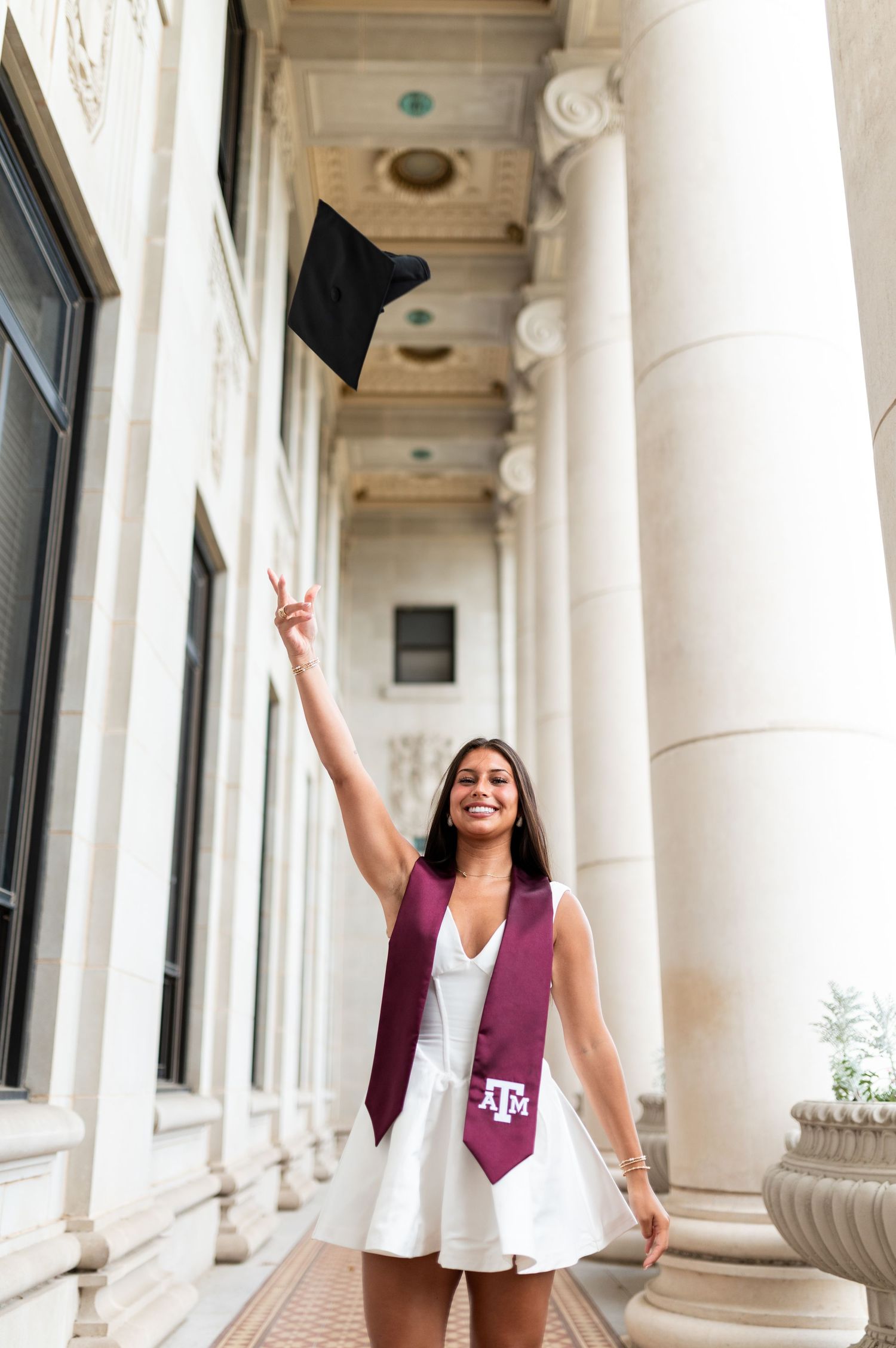 Texas A&M graduate in white dress tossing cap in the air outdoors. Grad Photos at the Admin building